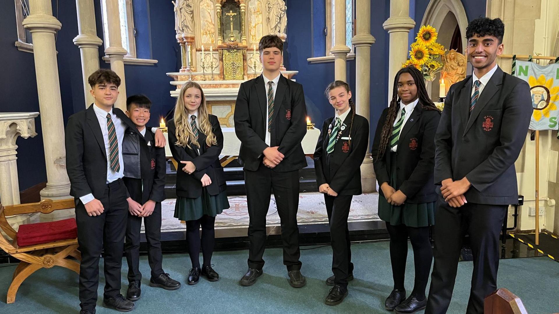 Pupils wearing school uniform inside a church with an altar behind them. A brass ornament of a cross and candles are on the altar.