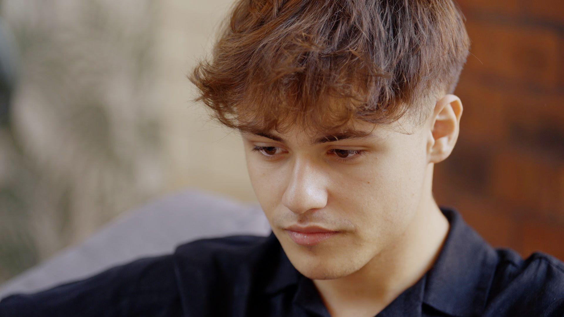 A close-up of Cai, a young man wearing a black shirt, looking pensive with his eyes cast downward