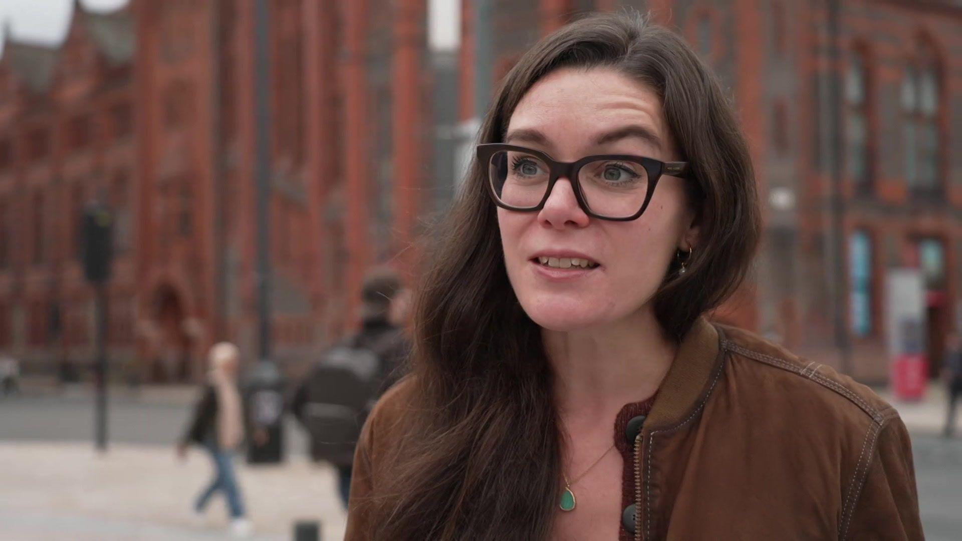 Dr Clarissa Giebel has long dark hair and is wearing dark framed glasses and a brown jacket. She is standing outside the University of Liverpool.