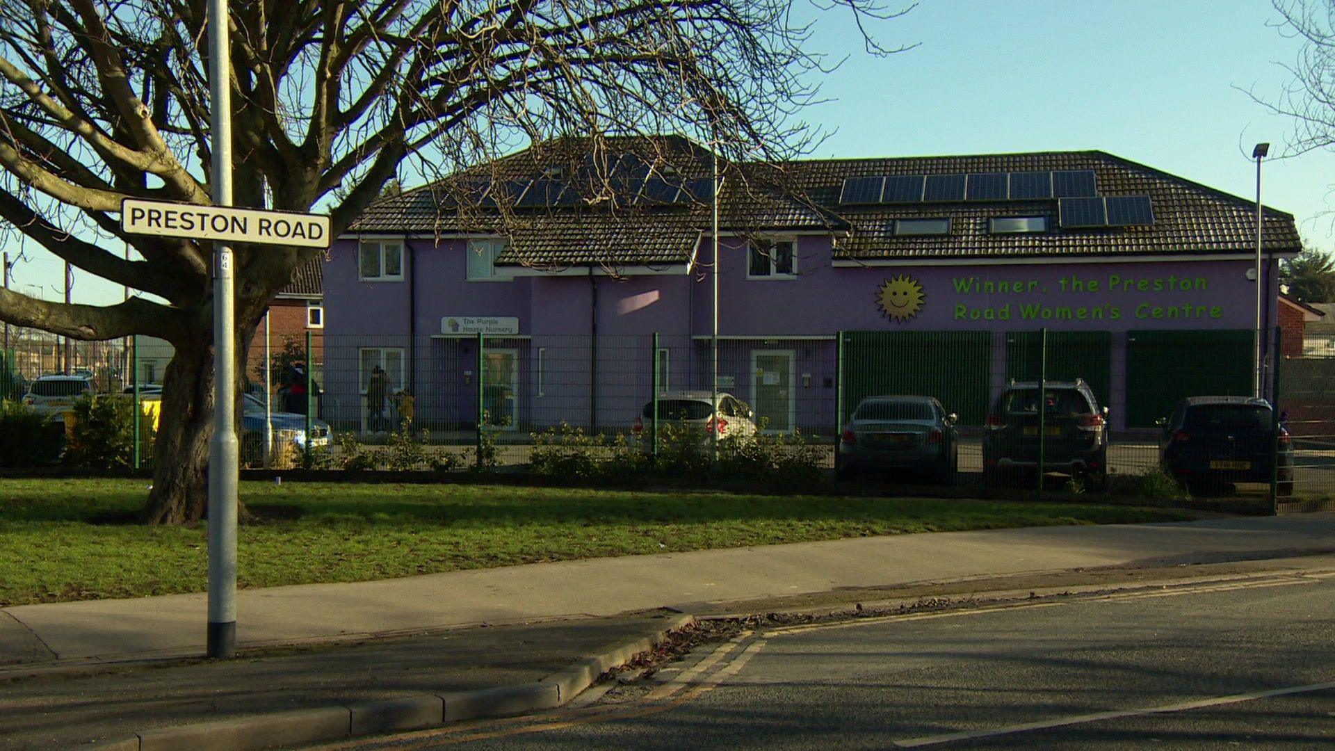 A building painted purple, with solar panels on the roof. It has a green fence around and the picture has been taken from across a road.