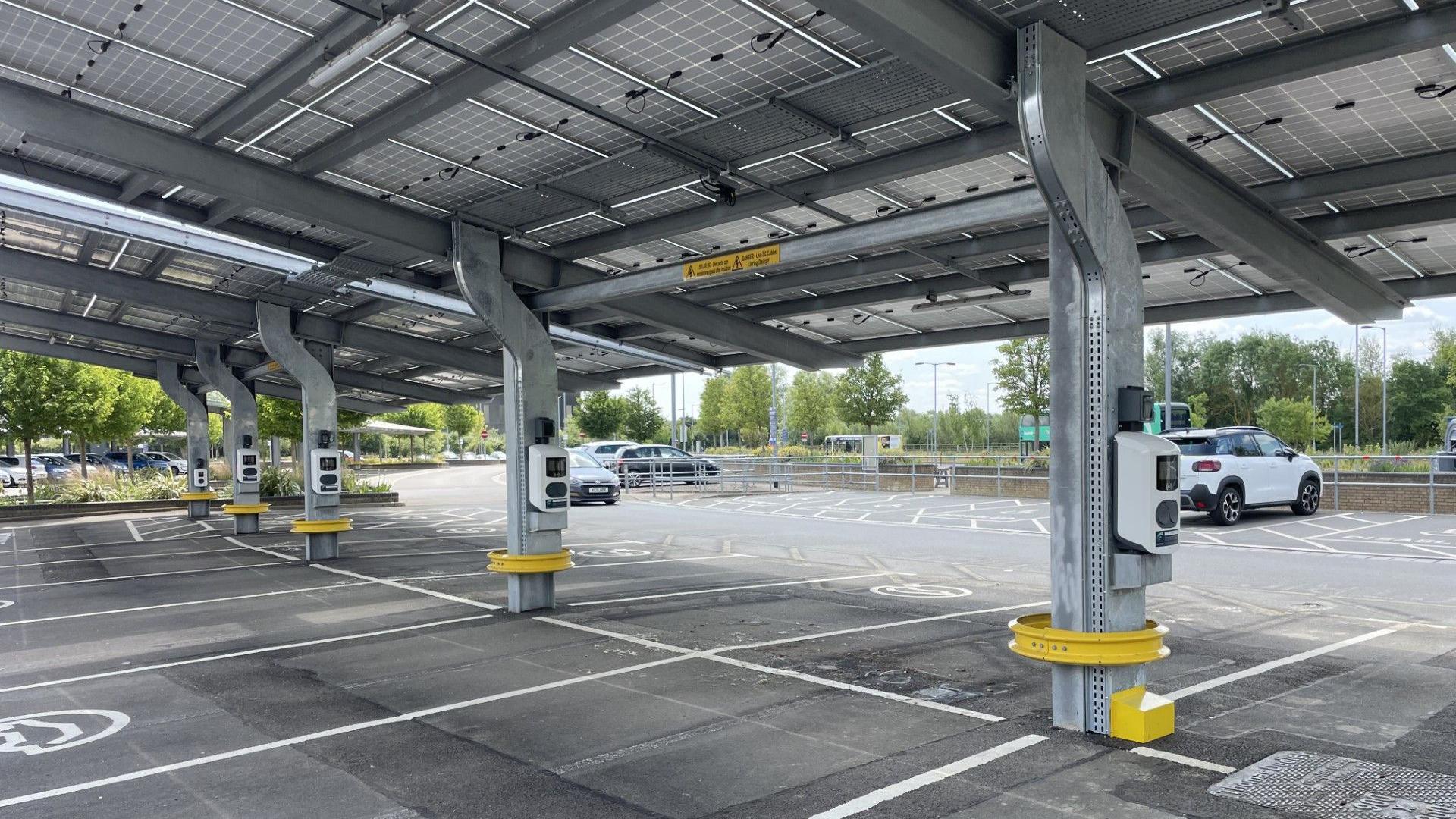 Electric vehicle charging points are installed on columns in a car park. There are solar panels on the roof above.