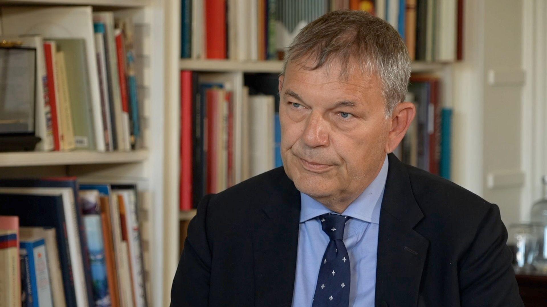 Philippe Lazzarini sits in front of a white bookshelf. He has greying hair and wears a blue shirt and dark jacket. He also wears a navy tie with small grey embellishments.