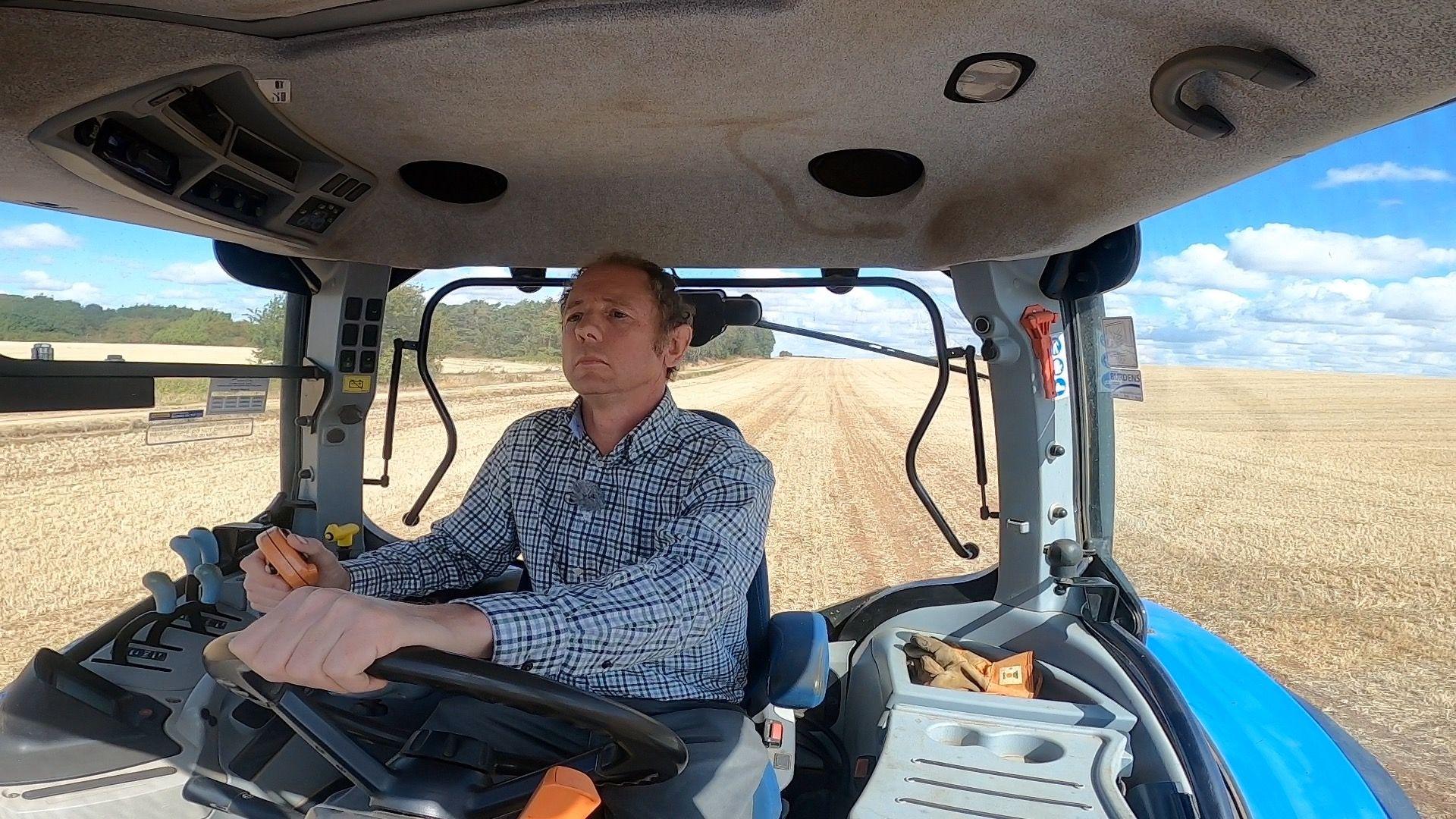 a picture of joe inside the cab of a blue tractor. In the background we can see the fields and skyline.
