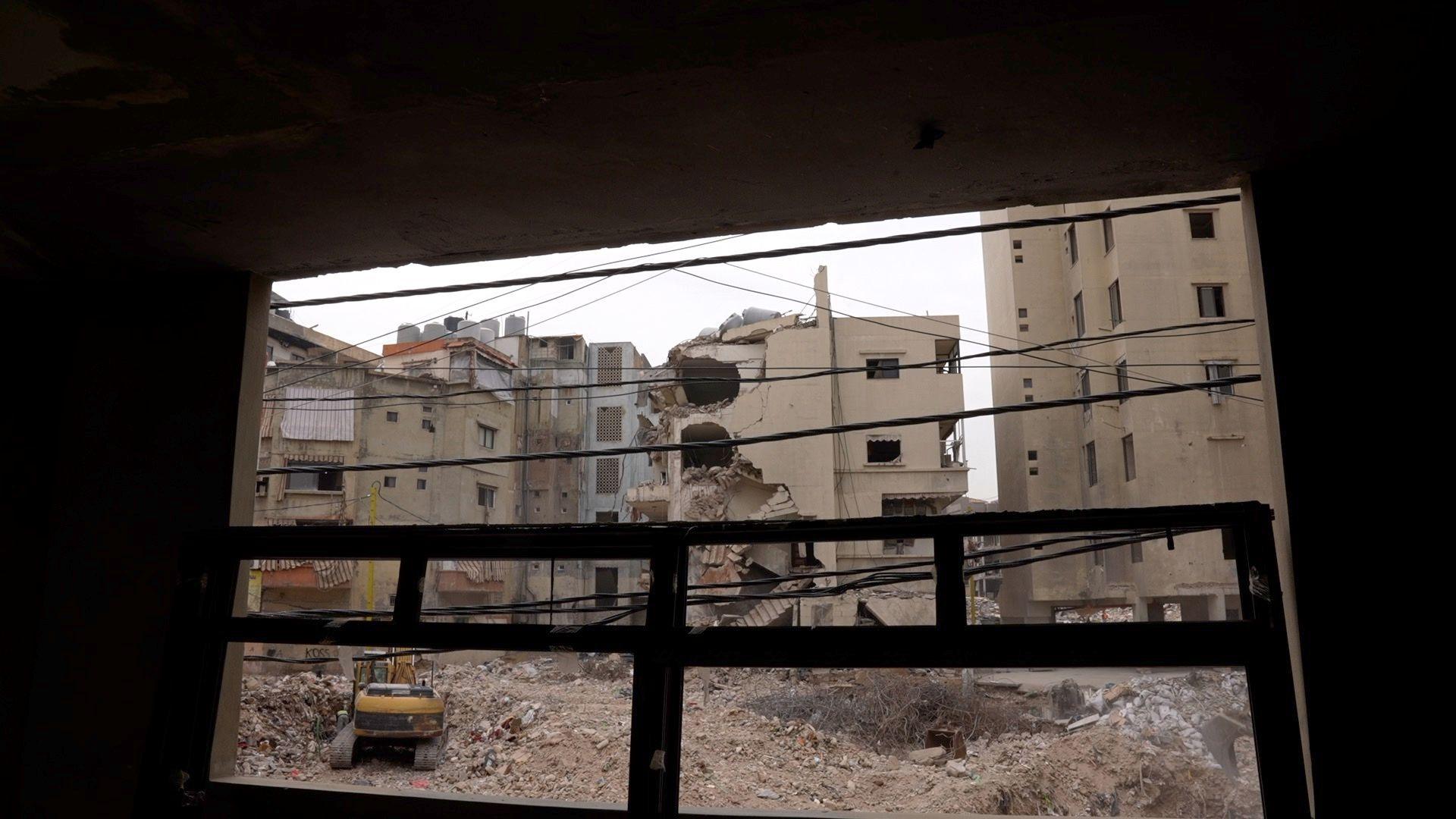 Standing inside a building of Burj High School looking out, a heavily damaged multi-story building can be seen, as well as large piles of debris and a yellow bulldozer.
