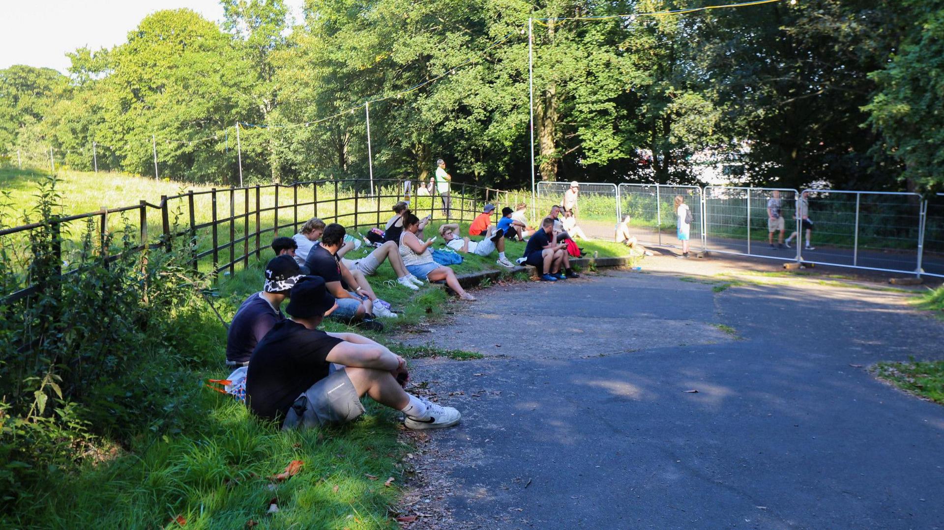 A number of Oasis fans in T-shirts, shorts and trainers sit in the shade of a wooded area alongside a path, which has been cordoned off with metal wire fencing, separating fans with Oasis tickets from those without