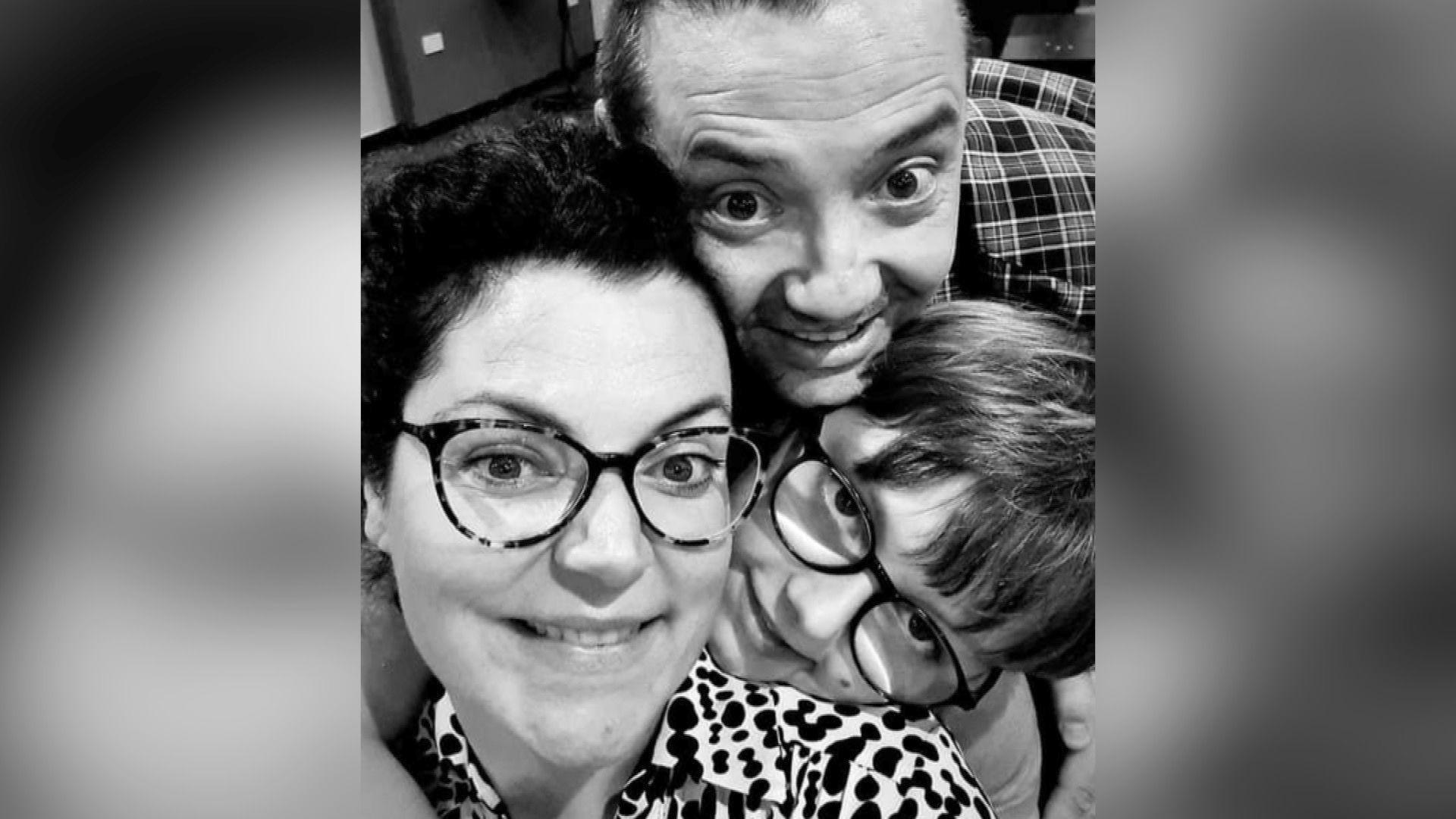 A black-and-white photo of Ashleigh Le Boutillier, a woman with dark hair and glasses, being hugged by Dean Lowe, a man with dark hair and Charlie Lowe, a young boy with glasses.