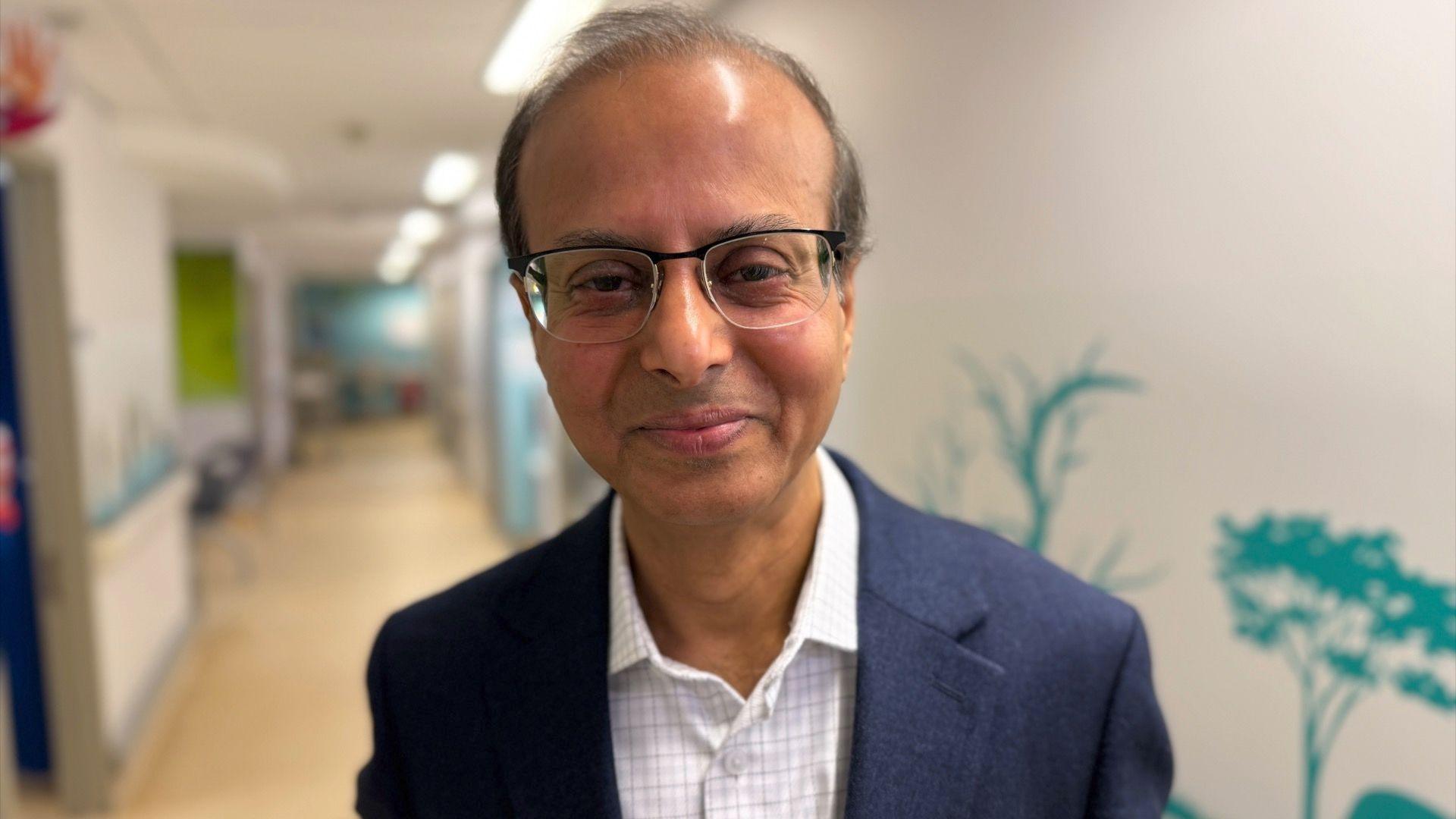 Dr Manas Datta wears a white shirt with light checked pattern, and a navy blue suit jacket over the top. He is stood in a hospital corridor with white walls behind him and turqouise-coloured stencils of plants on the walls. Dr Datta has dark thinning hair and wears spectacles. He is smiling at the camera.