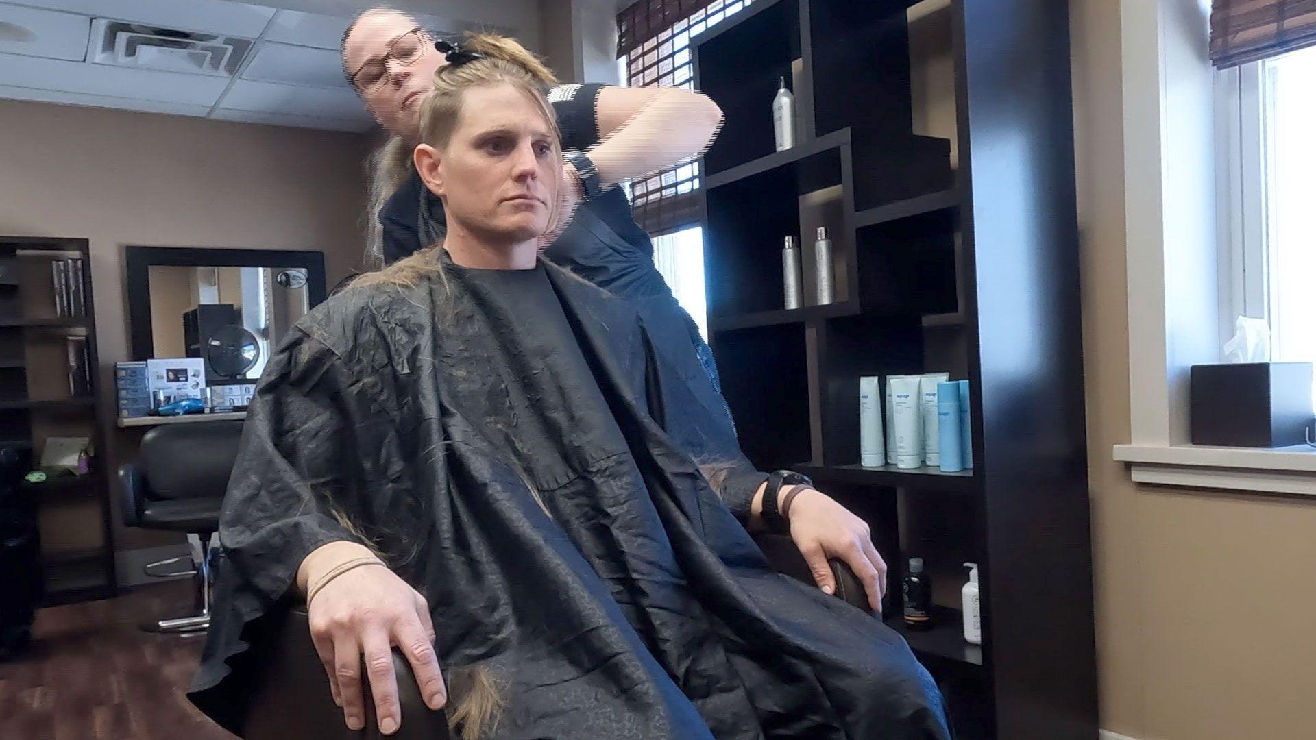 Kara sits in a chair in a hairdressing salon, wearing a black cape over her clothes, while a hairdresser cuts her hair.