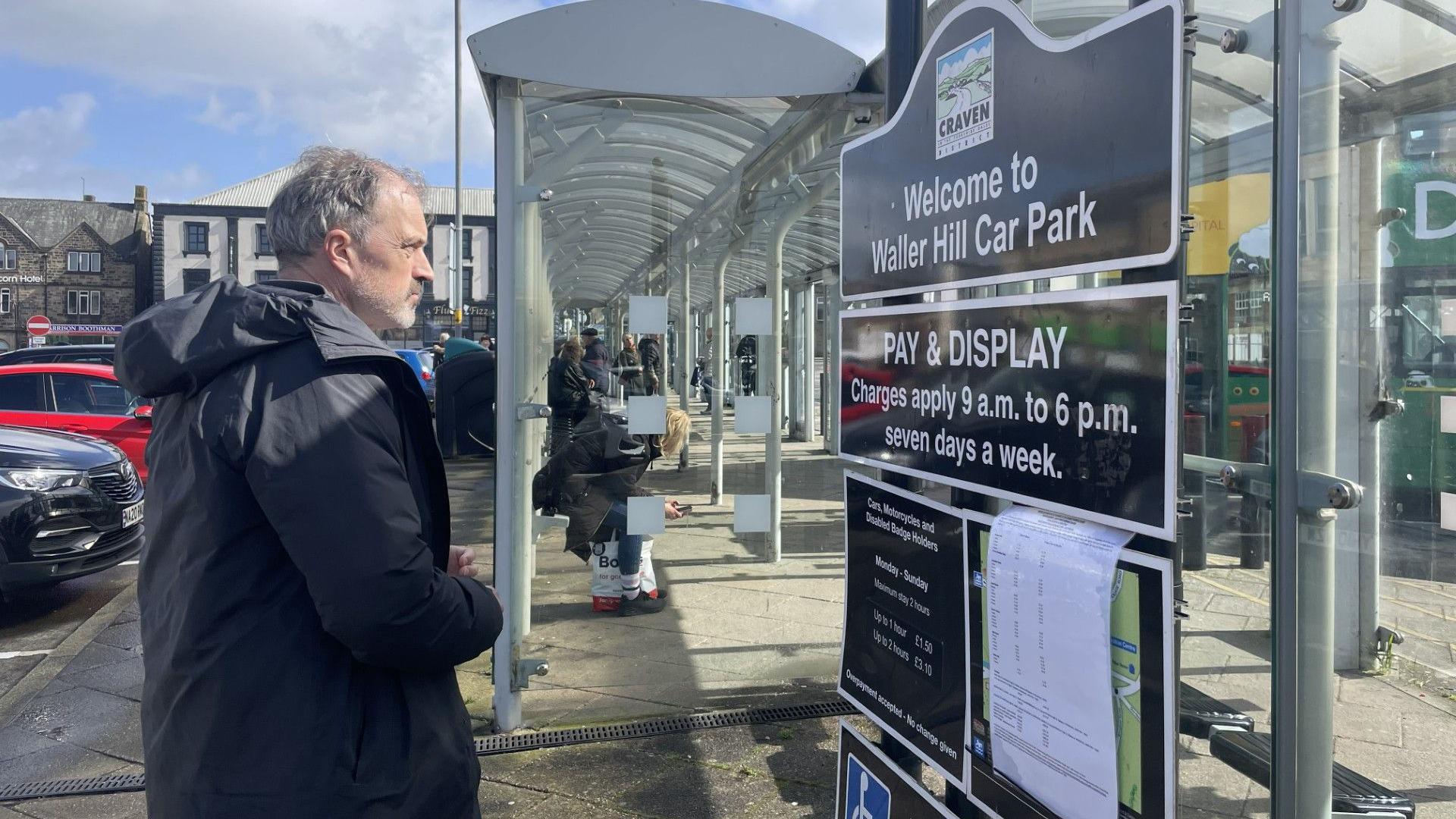 MP Sir Julian Smith is wearing a dark-coloured coat and is reading a sign at Skipton bus station that says 'Welcome to Waller Hill Car Park' with a pay and display sign below. Behind him is a long bus shelter with people waiting for buses and some parked cars.