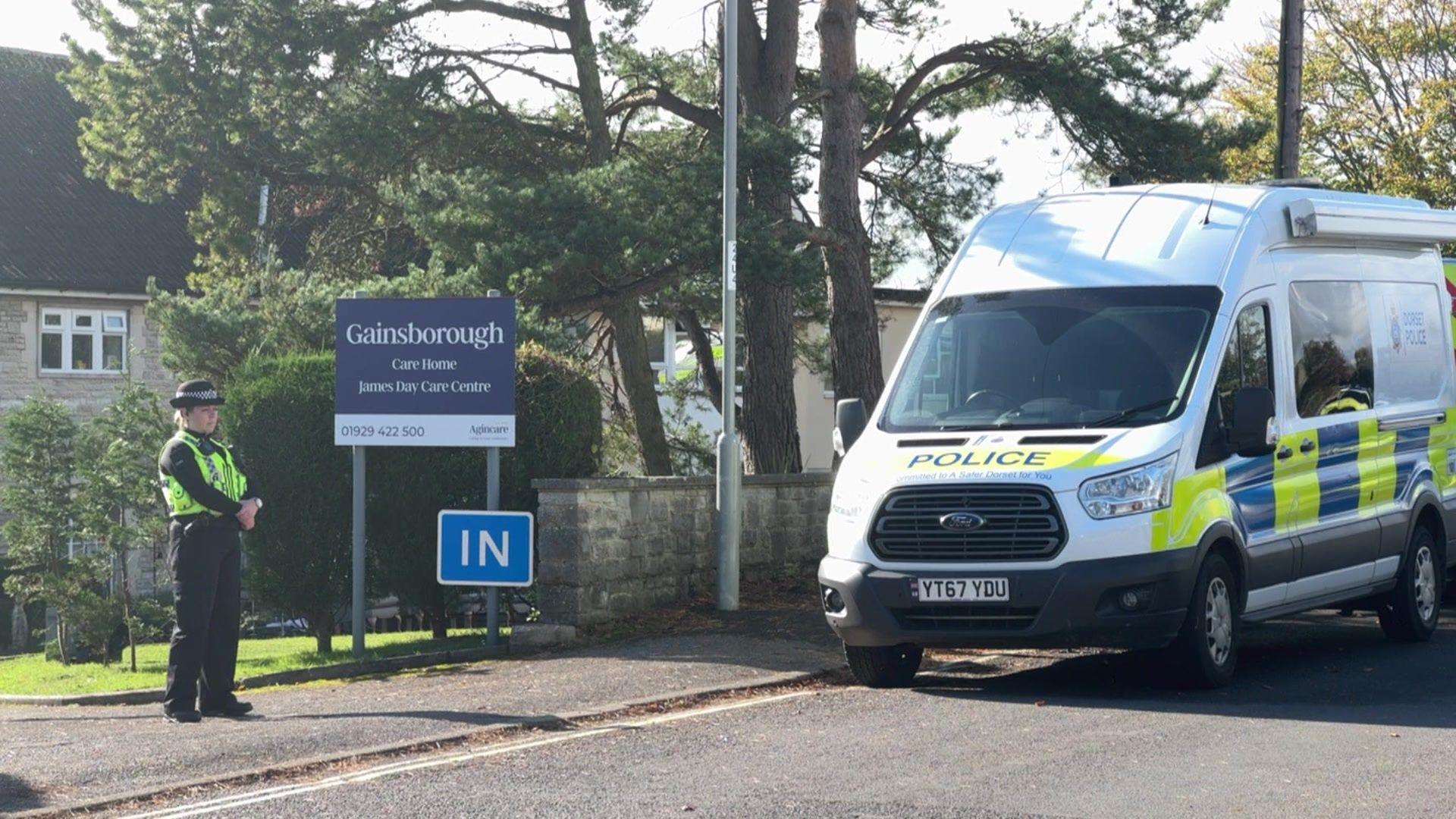 A female police officer stands at the entrance to Gainsborough Care Home. A marked police van is parked on the roadside. The two-storey building can just be seen behind some trees and a blue sign saying 'Gainsborough Care Home'.