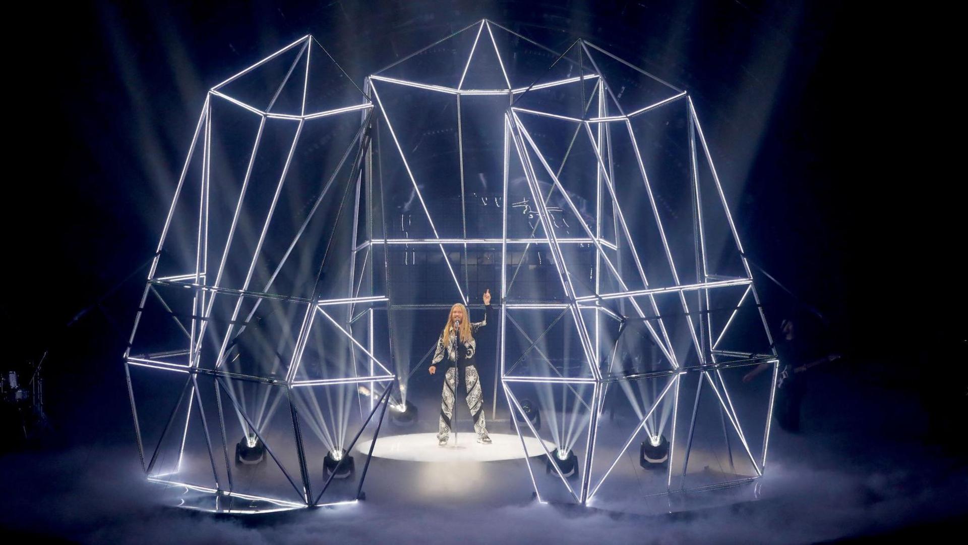 Ryder is wearing the jumpsuit on stage during his Eurovision performance. He is surrounded by a metal structure that is lit up with white lights.