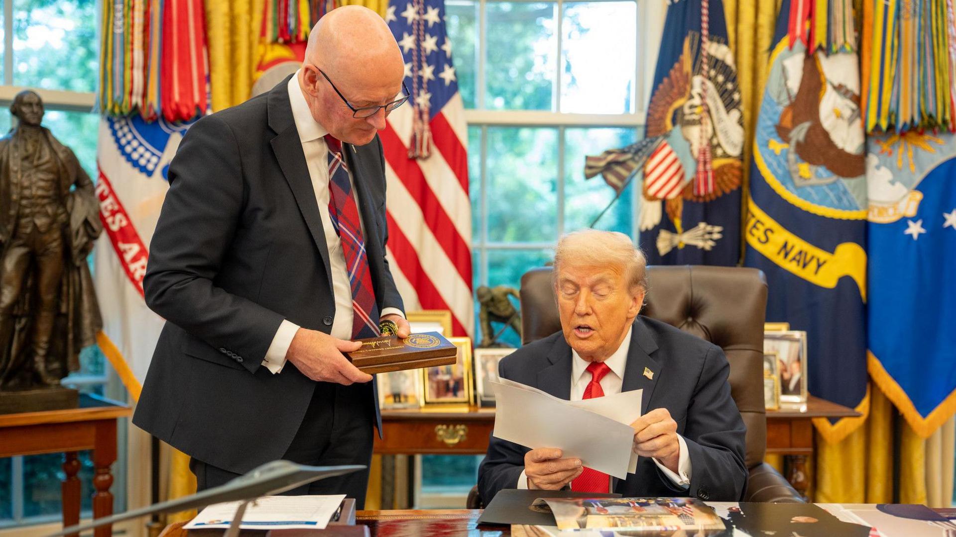 John Swinney and Donald Trump in the Oval Office. Swinney is holding a wooden plaque and Trump is holding pieces of paper in his hands.