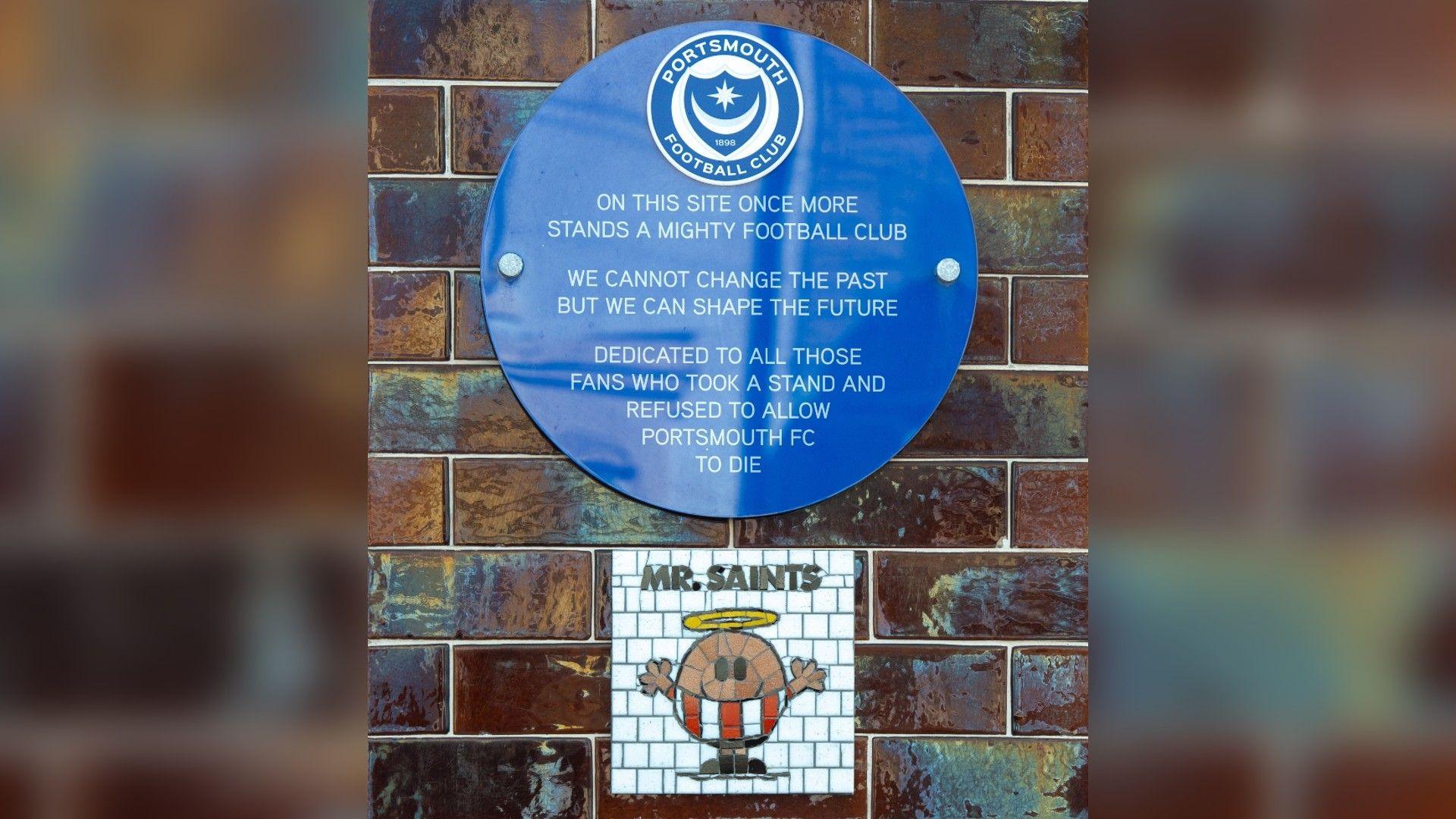 A blue plaque which shows the Portsmouth Football Club crest above text which reads "on this site once more stands a mighty football club. We cannot change the past but we can shape the future. Dedicated to all those fans who took a stand and refused to allow Portsmouth FC to die" underneath the plaque is a white mosaic with a mr man character dressed in a southampton fc kit, it says "mr saints" at the top