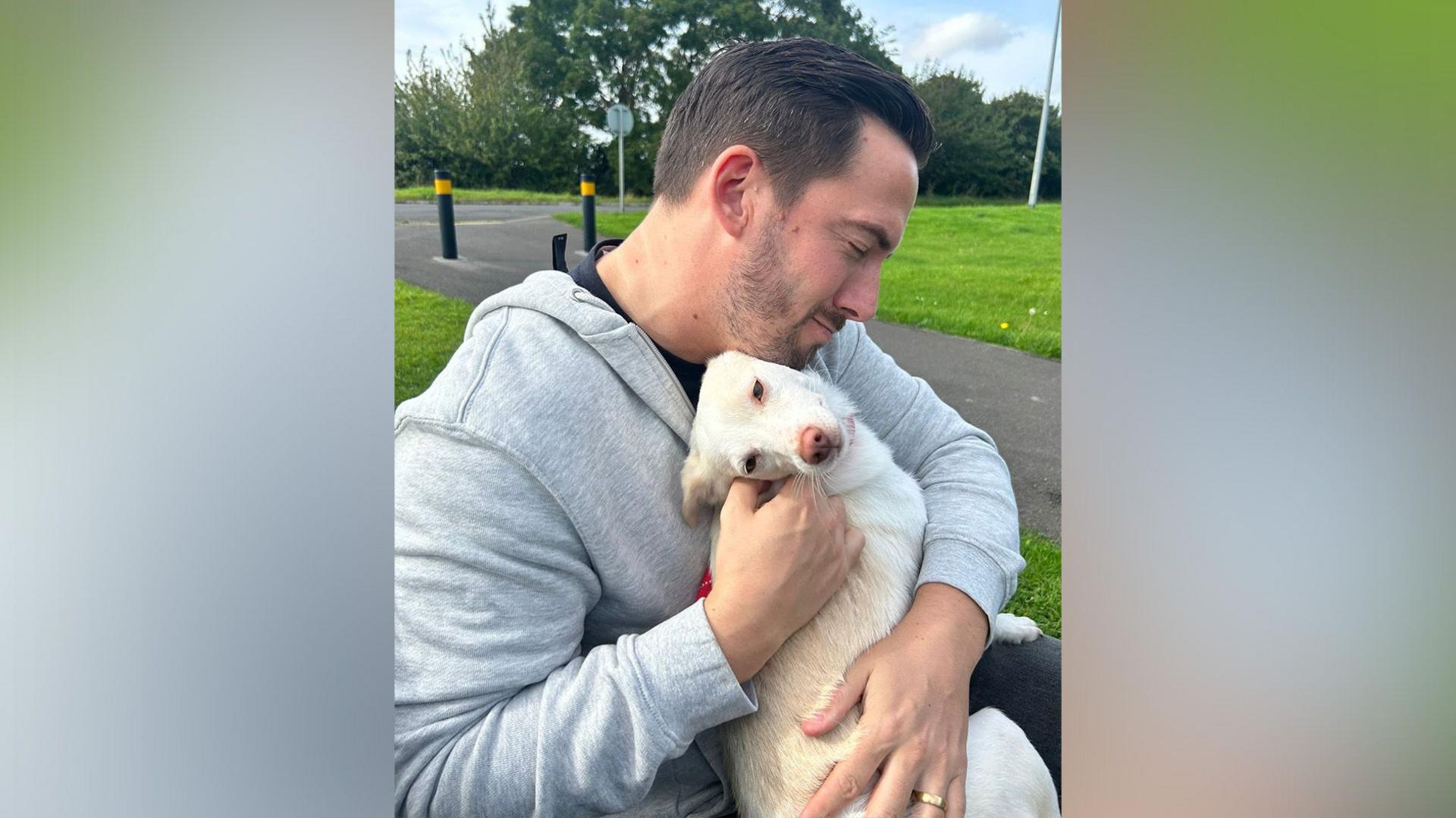 Andy Marriott sits on a bench cuddling Laika, their much-loved family pet. Laika is leaning back and nuzzling against Mr Marriott as she is stroked.