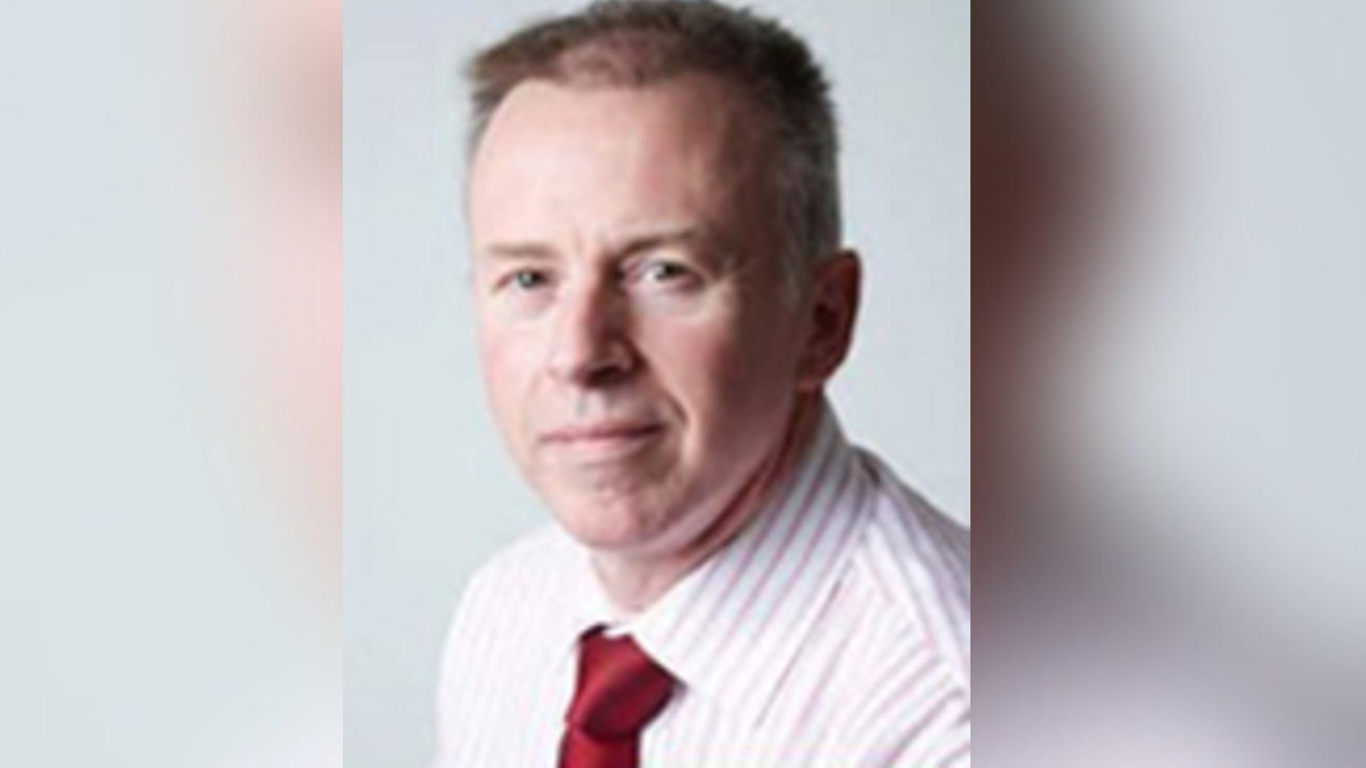 A short-haired man with a pink and white stripy shirt and red tie looks at the camera.