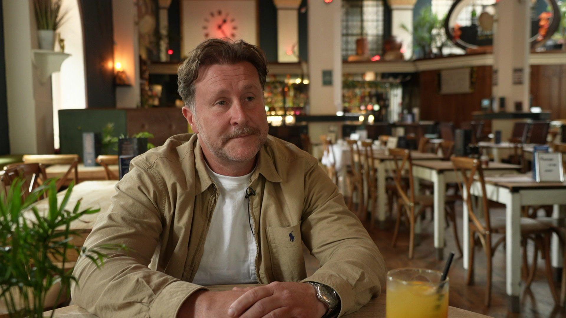 A man is pictured sat at a table inside of a bar. He is wearing a silver watch on his left wrist and is wearing a white shirt with a cream jacket. His hair is slicked back and he has a moustache. 