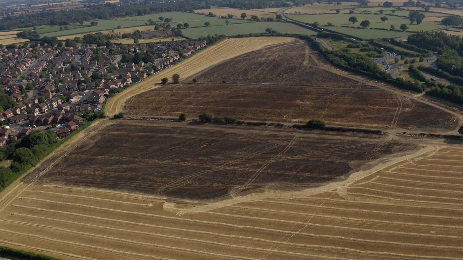Large fields. A huge part of the fields are burnt and darkened. A road is to one side of the fields but houses are to the other.
