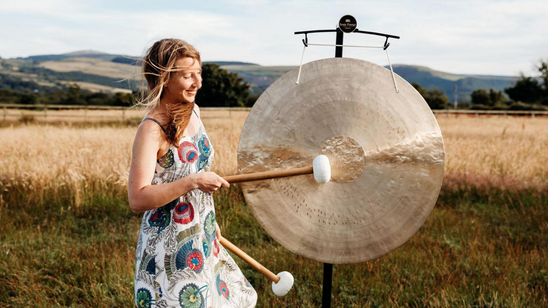 Amy Howse banging a large golden coloured gong while standing in front of a field with golden crops. She has long brown hair and is wearing a white summer dress with coloured patterns on it.