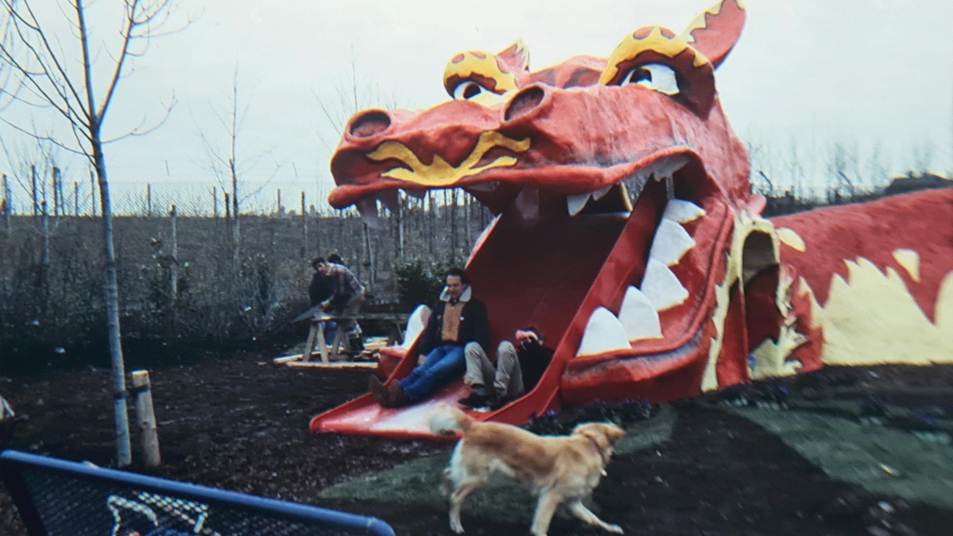 A young Theo Gay Anderson, seen lying on his back, and Blue Peter presenter Simon Groom finish riding down the schoolboy-designed dragon slide. The dragon is a large red-and-yellow fibreglass construction. Blue Peter's pet dog Goldie is running in front of the slide.