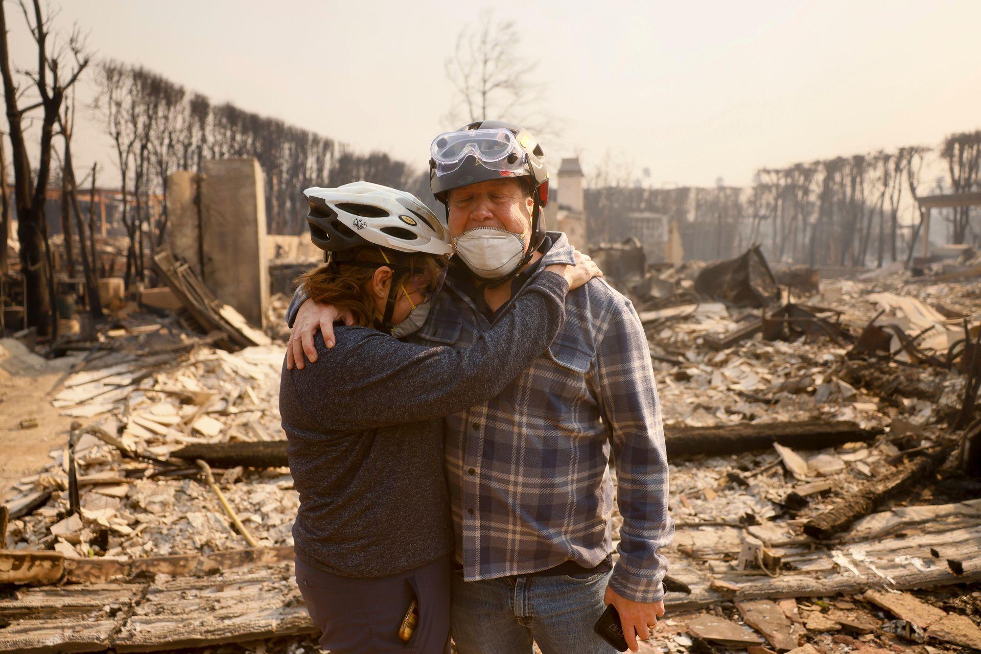 A husband and wife embrace in their fire-ravaged neighborhood, in front of burnt rubble, both wearing helmets and protective face masks, after the Palisades Fire swept through in the Pacific Palisades neighborhood of Los Angeles on Wednesday.