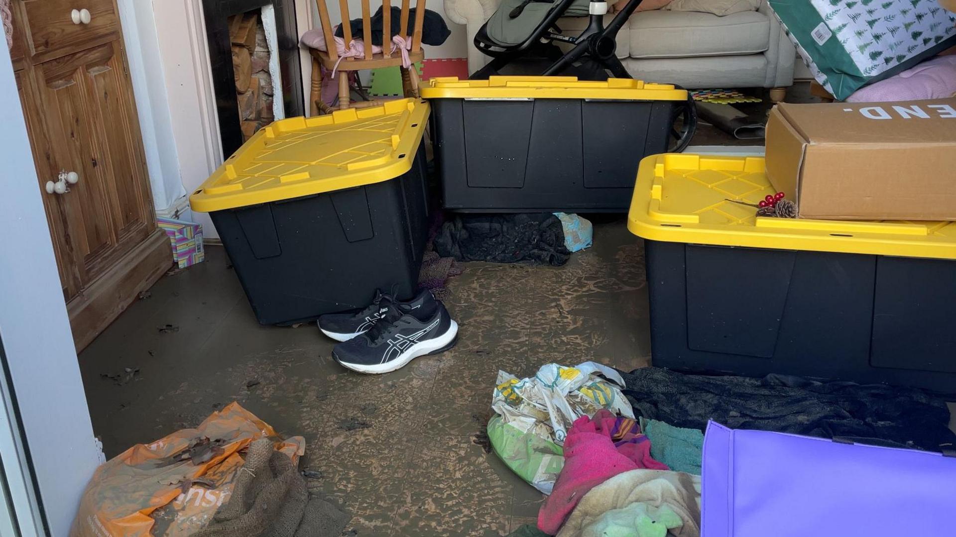 Boxes with yellow lids and a pair of trainers on a muddy floor. Other items can be seen piled up in the background.