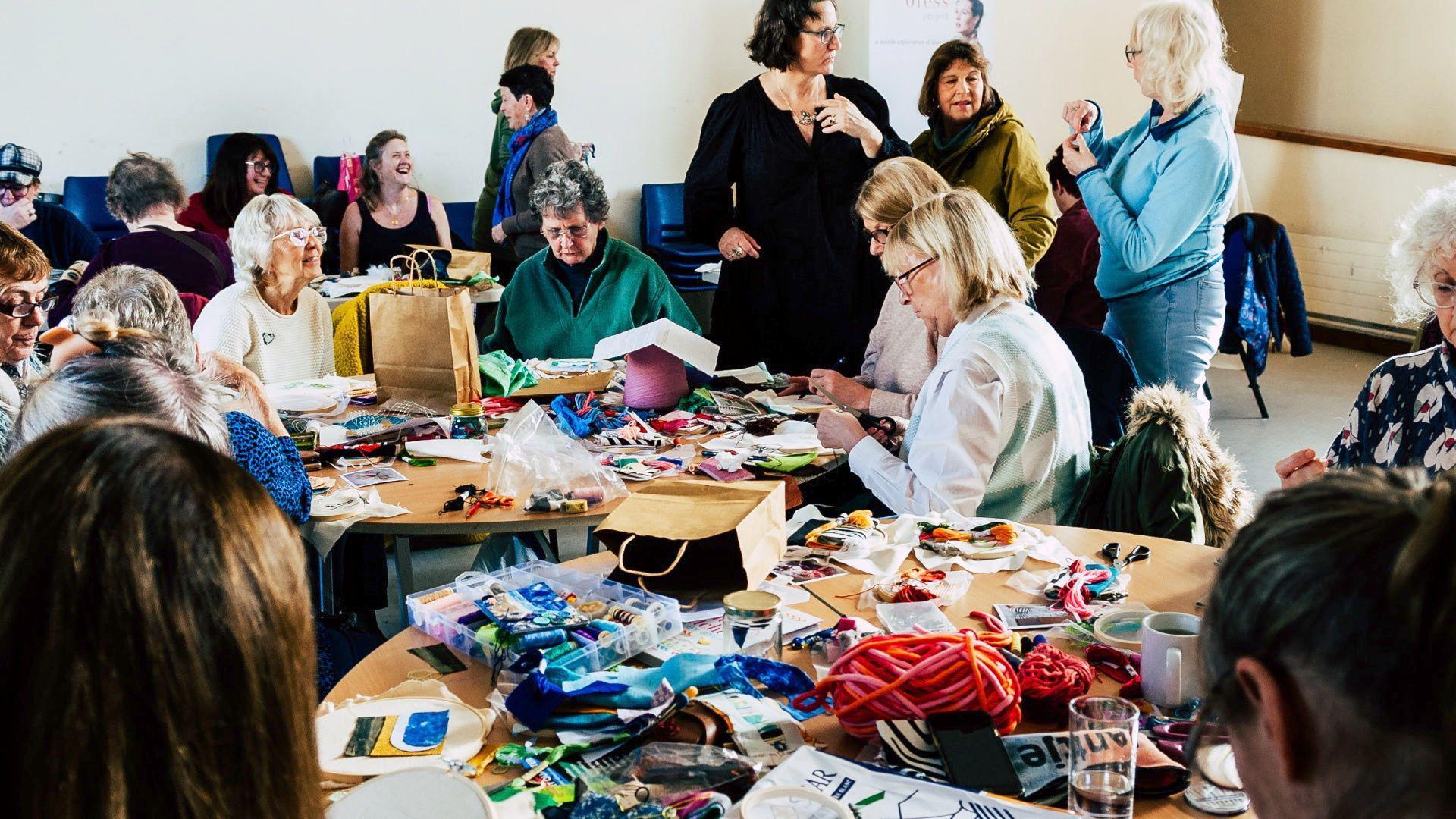 Dozens of women are sat around a number of tables sewing and talking. Some are also standing chatting. There are sewing materials all over the tables, many are smiling while they work.