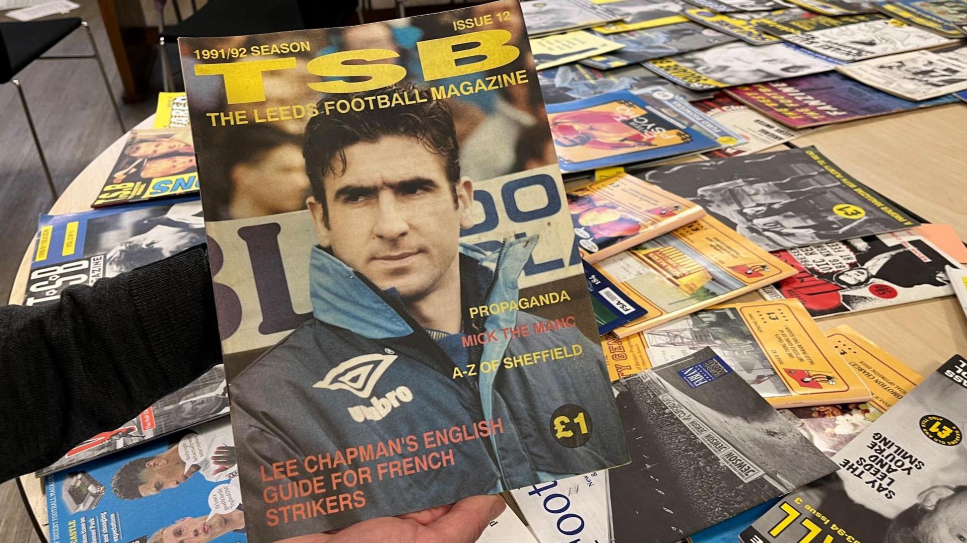 A picture of a person holding a fanzine with the words 'TSB The Leeds Football Magazine' on the front. In the background are dozens of other fanzines spread out on a table. 