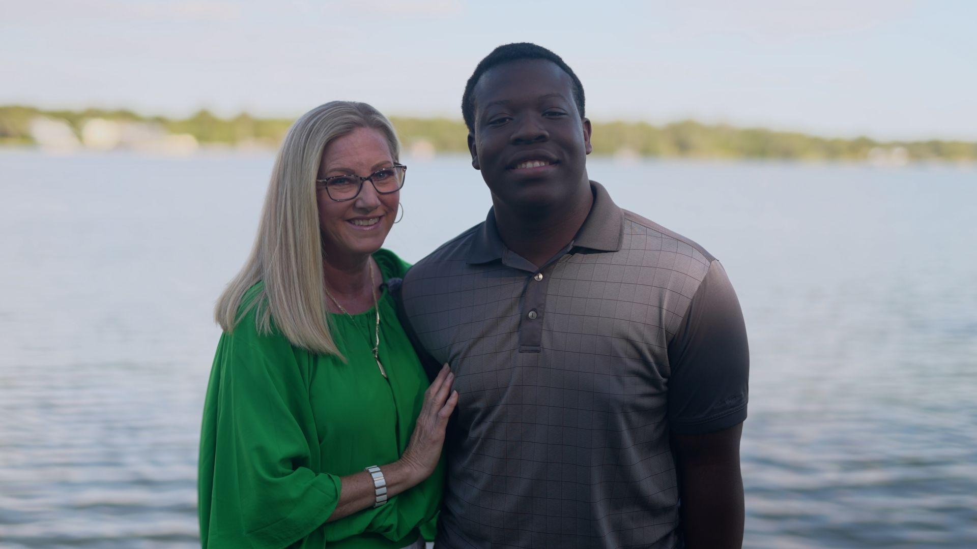 Stacey and Marven facing the camera in front of a lake. Both are smiling, Marven is wearing a grey, collared T-shirt, Stacey has long blond hair and glasses and is wearing a bright green gathered top.