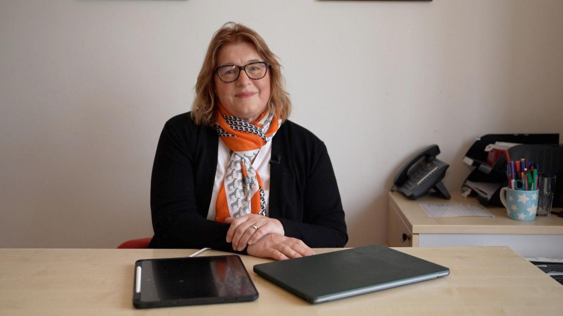 Dr Georgie Siggers sits behind a desk. She is wearing glasses, a black cardigan and white blouse and an orange, white and blue scarf.