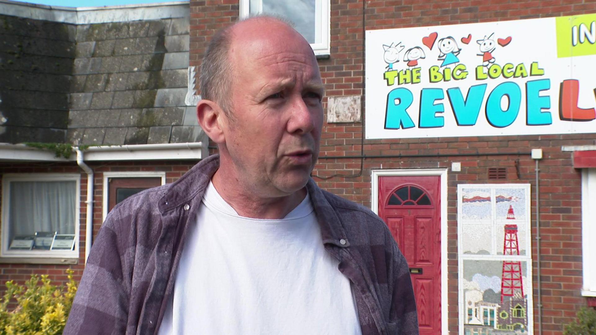 Simon Lawton, wearing a white t-shirt under a cheque patterned fleece, speaks to the camera in front of a building adorned with a sign for Revoelution