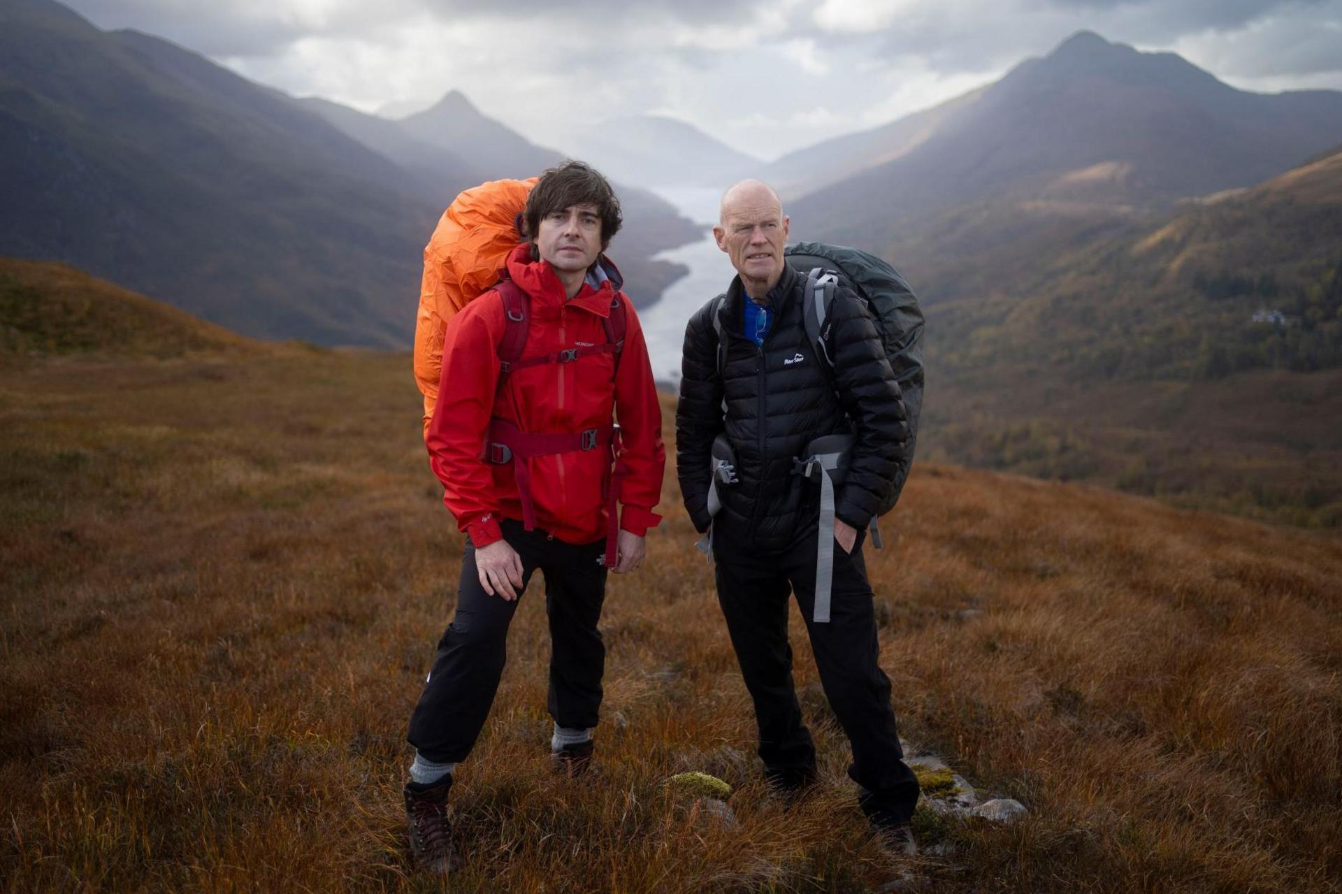 Danny and Phil are wearing outdoor clothing. They are carrying rucksacks. The men are standing on a hill with loch and mountains behind them.