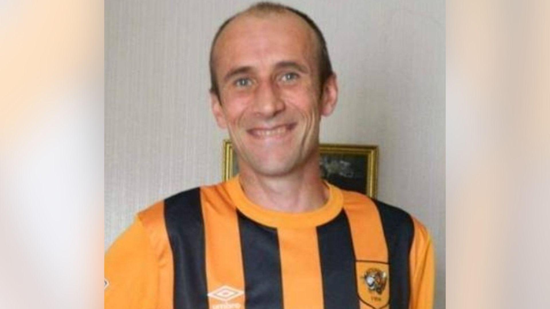 Christopher King, smiling and wearing a black and amber striped Hull City football shirt. He is standing in front of a painting. Mr King has blue eyes and balding dark hair.