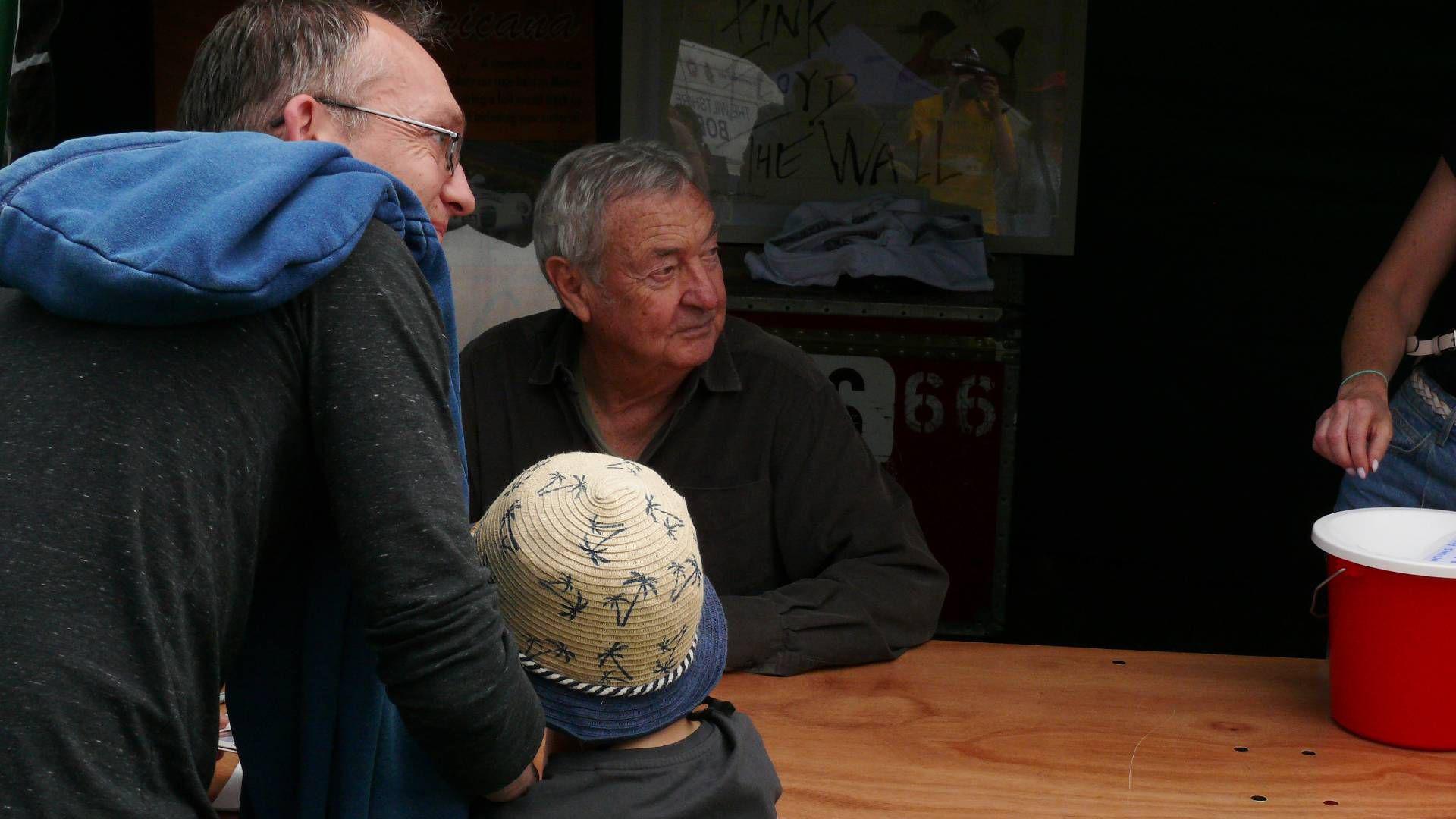 Nick Mason sitting at a wooden table and looking off into the distance. In front of him is a young boy and his father.