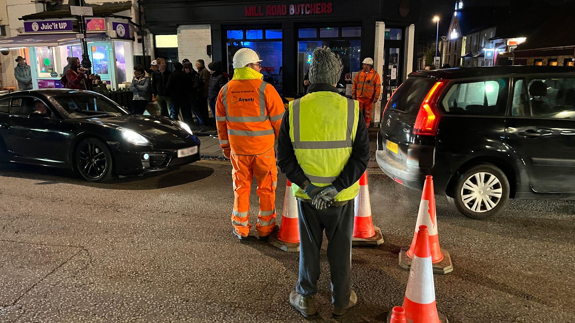 Two workmen can be seen and a person in a hi-vis vest are stood near a line of cones. Two cars are being let through the line of cones. People can be seen lining the road in the background. 