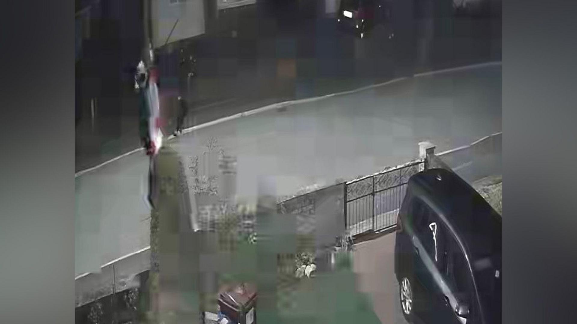 A distorted CCTV image showing a driveway with gates and a car parked in the bottom right hand corner. There appears to be someone climbing a lamppost where there is a red and white flag.