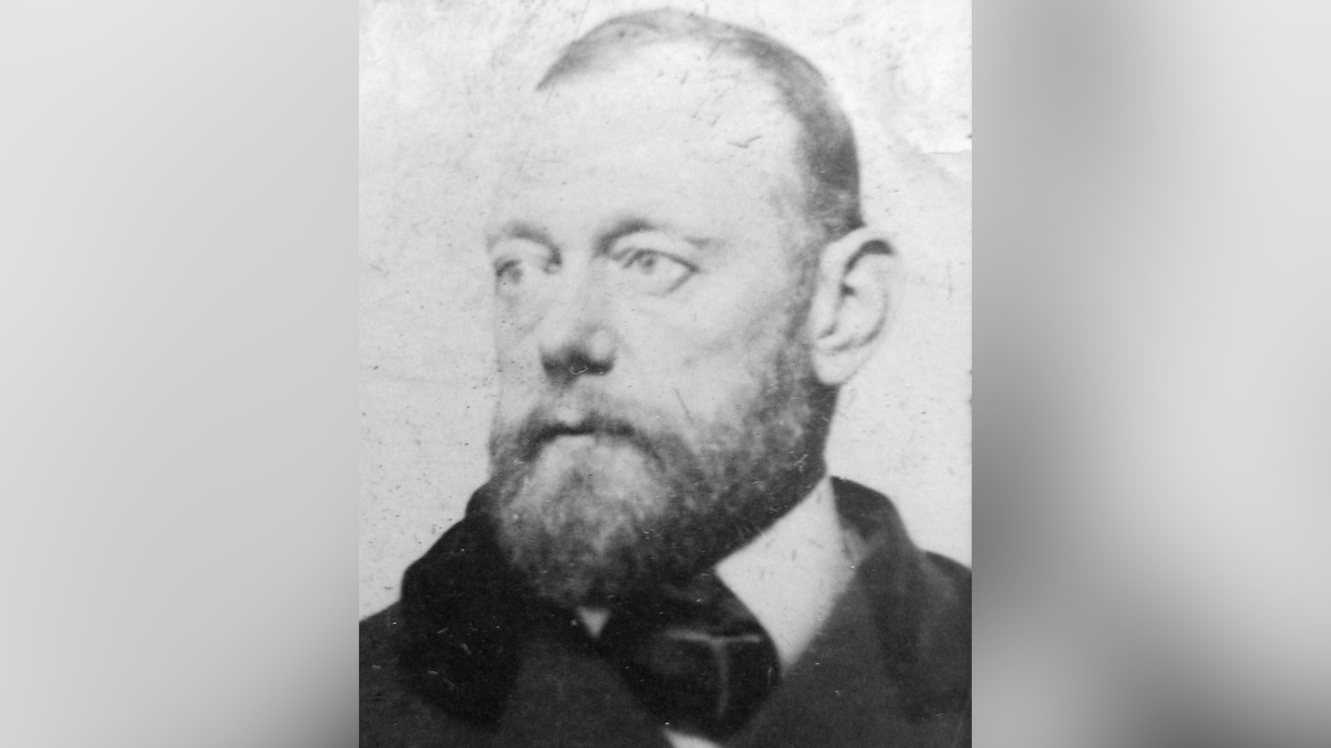A black and white photo of a bald man with a beard. He is wearing a suit and tie and a coat.