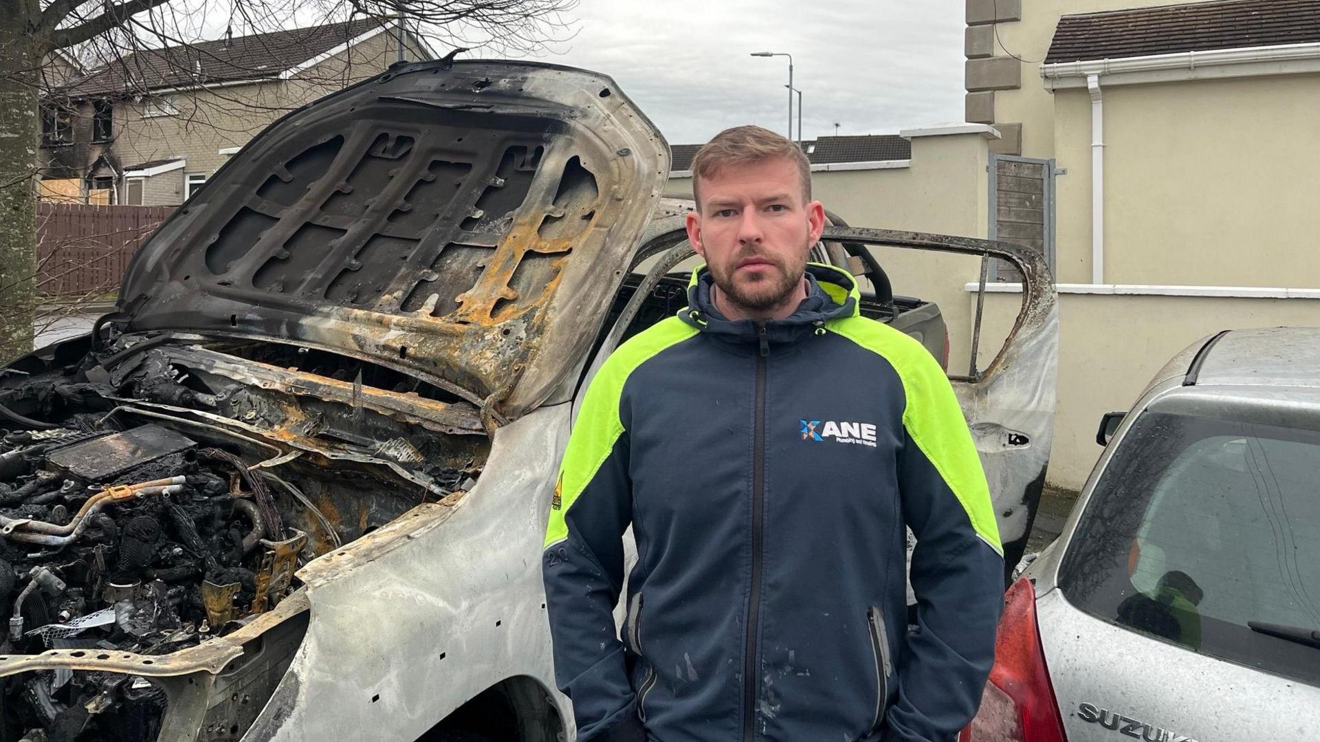 A man standing in front of a burnt-out car, he is wearing a yellow and blue navy. He has light brown hair and a beard.