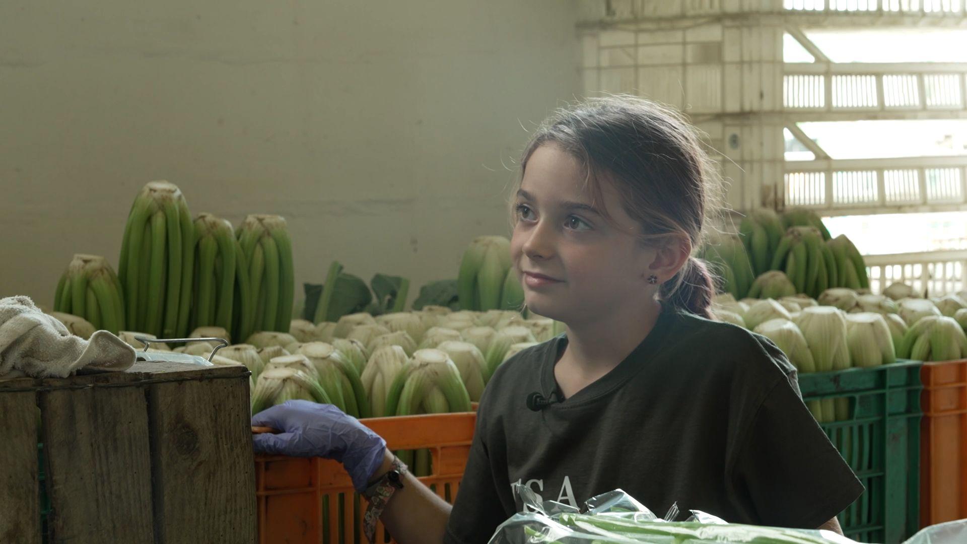 A girl standing in front of boxes of packed celery. The girl has a dark green t-shirt and blue plastic gloves.