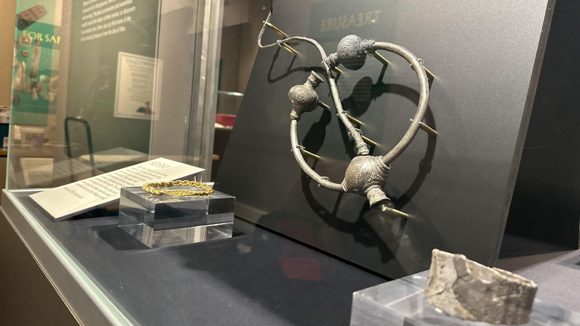 A large silver broach which is a circle with round decorated joins and a large pin through the middle. It is in a glass case with silver and gold bracelets.