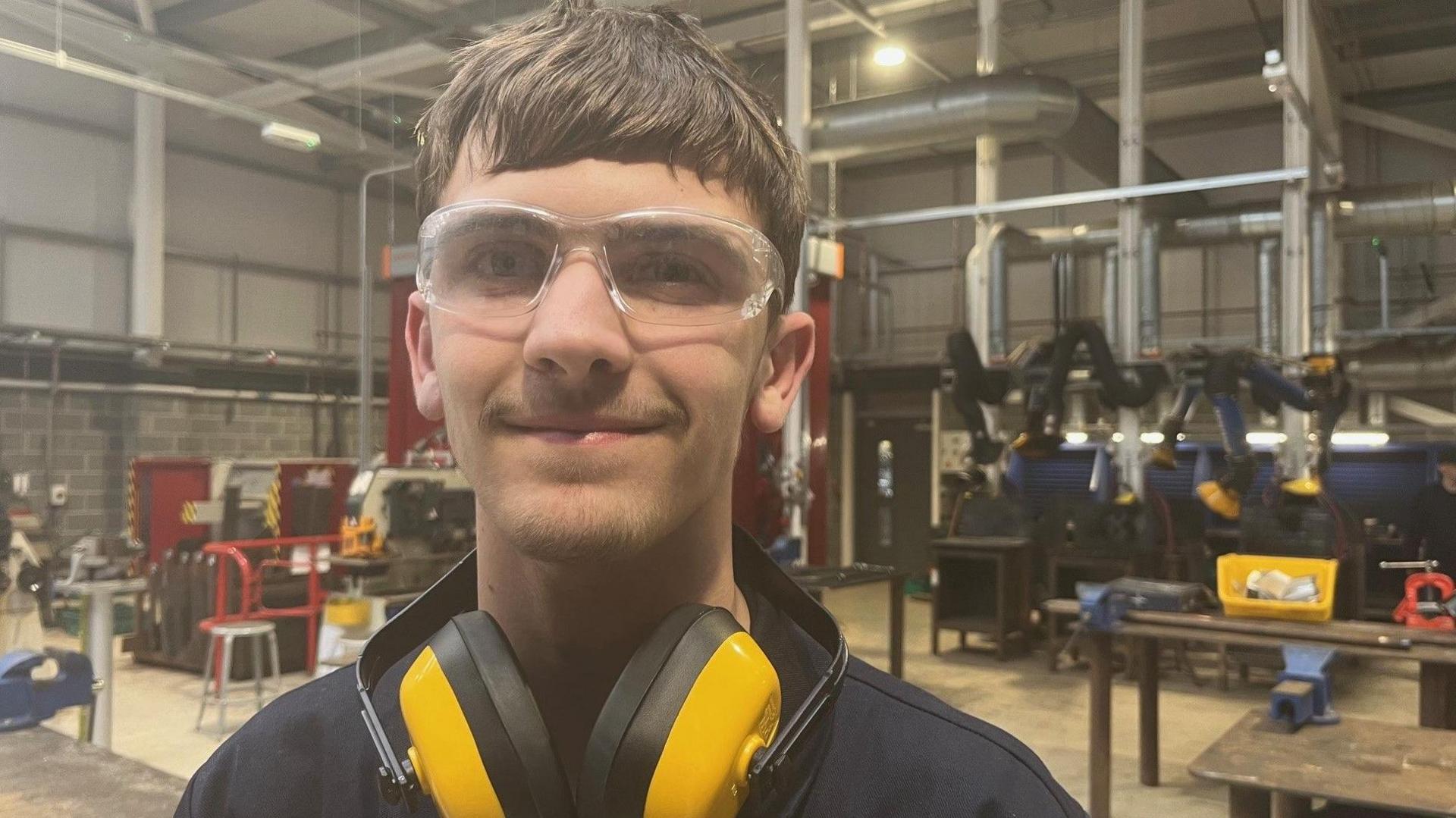 A young man with short brown hair smiles looking at the camera a he stands in a workshop. He is wearing blue coveralls and safety glasses. He has yellow ear defenders around his neck.