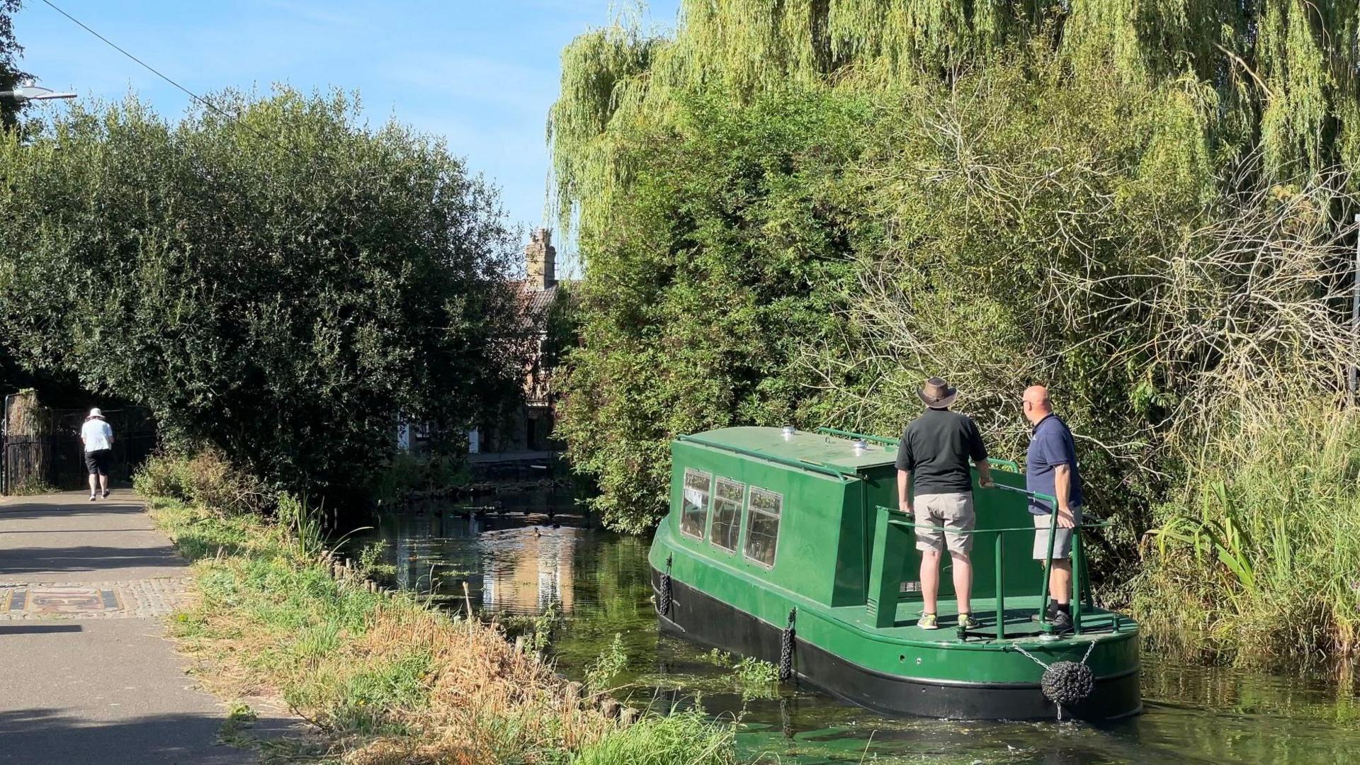 The green narrowboat sailing on the river, lined with trees