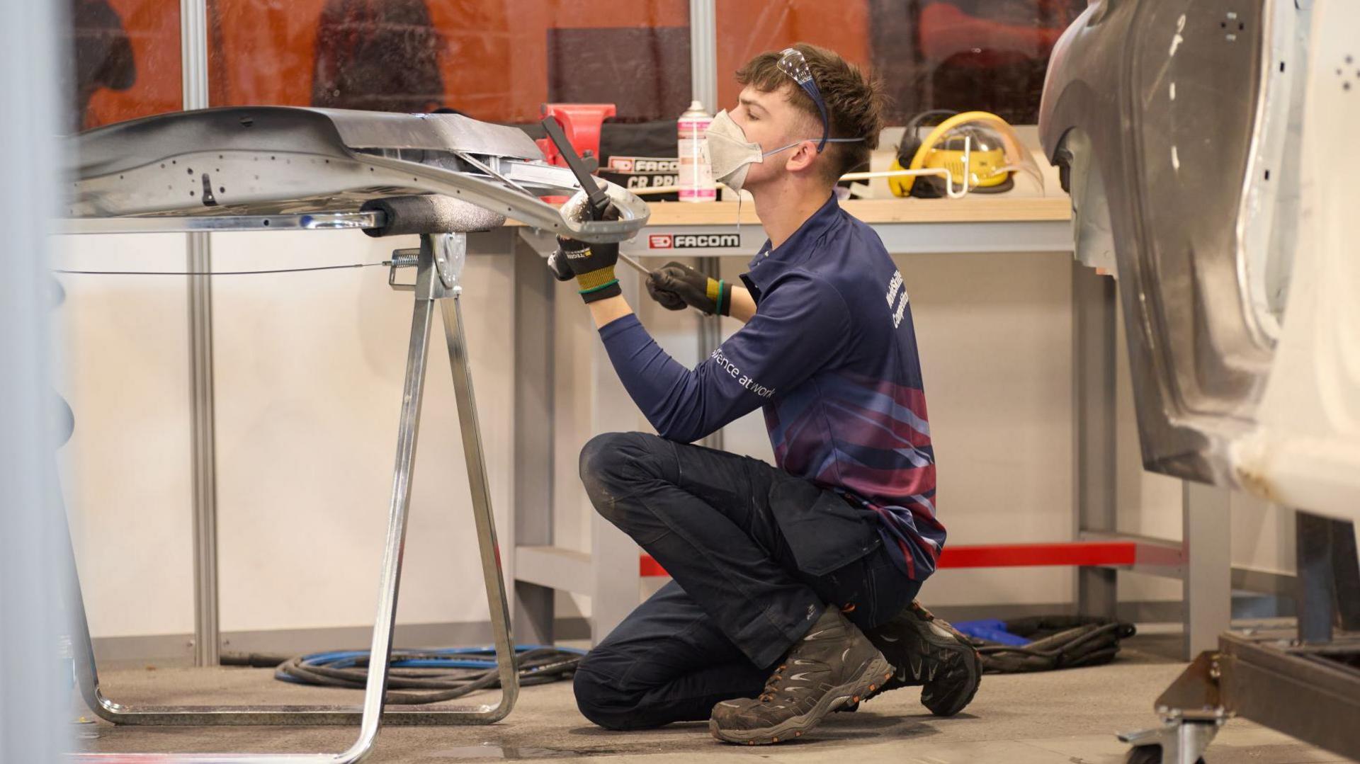 A man kneeling on the ground working on a car panel. He is wearing a navy long-sleeved polo shirt with red detailing, work trousers and brown boots. He has a pair of glasses on his head and is wearing a mask.