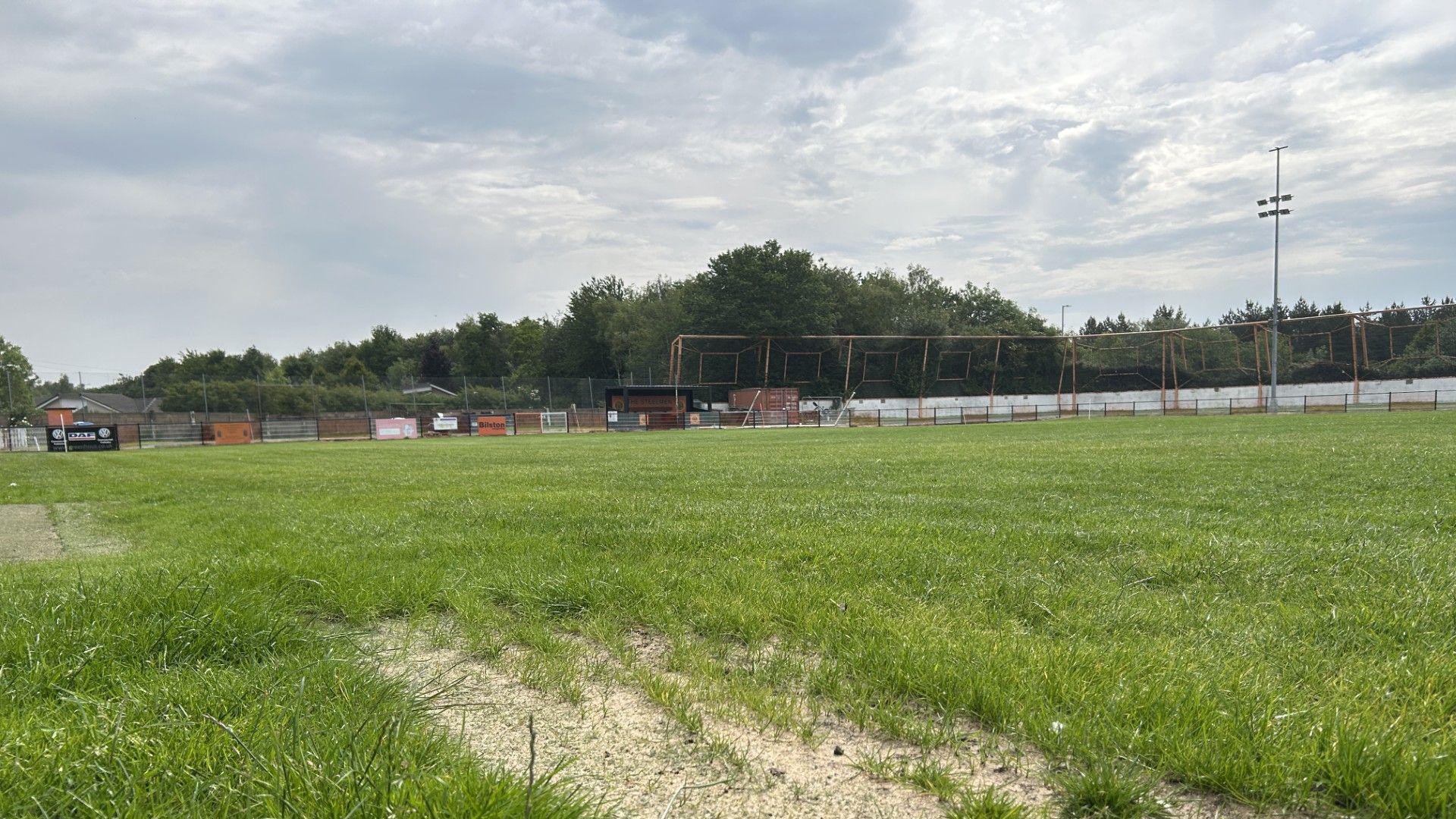The pitch of Bilston town. The grass is green but looks dry. It is an overcast day and we can see an orange fence and flood lights in the background.