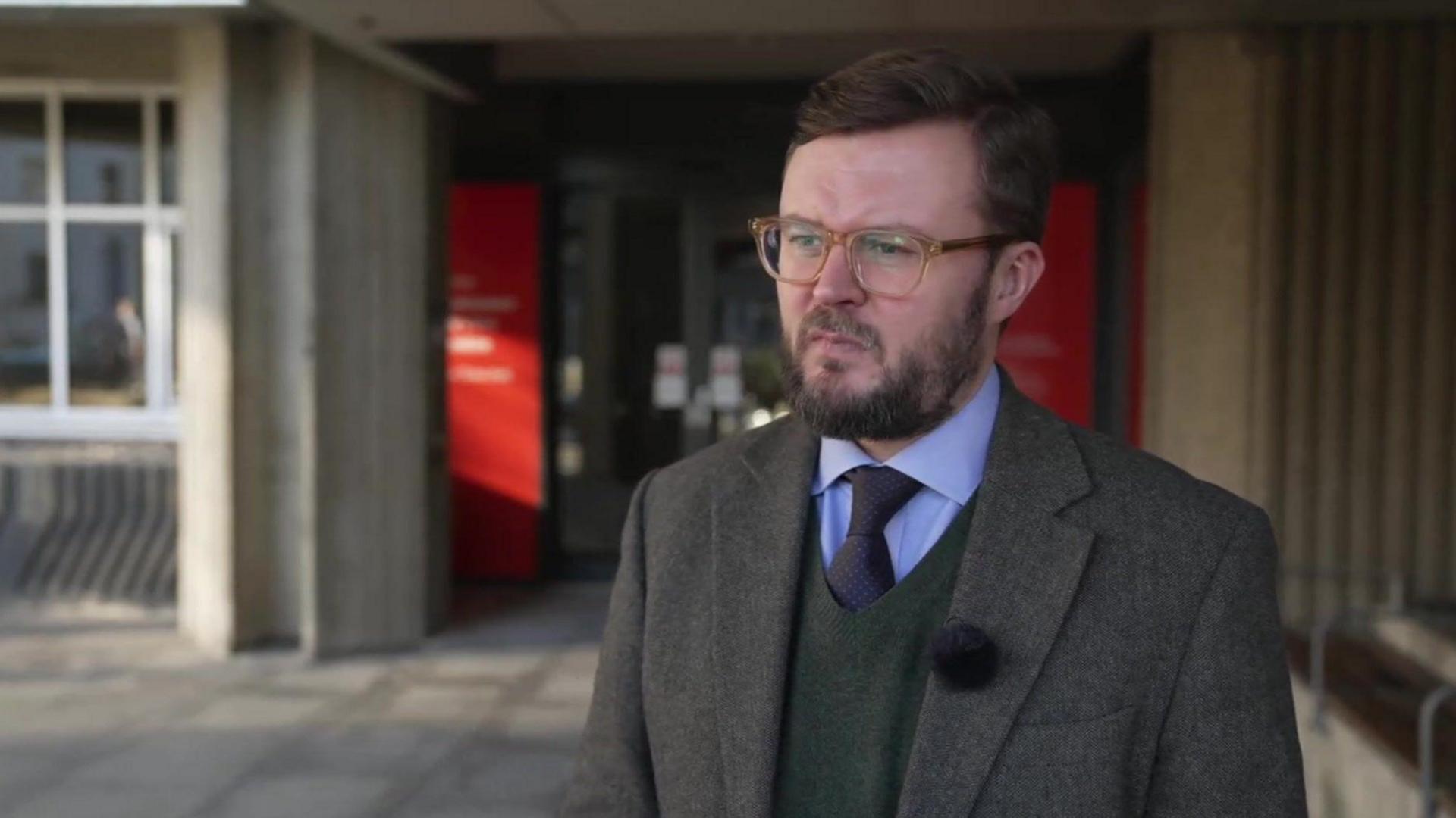 Jacob Taylor, being interviewed standing outside Hove Town Hall. He wears a dark grey jacket, green pullover, blue shirt and dark tie, has a beard and moustache and wears horn-rimmed glasses.