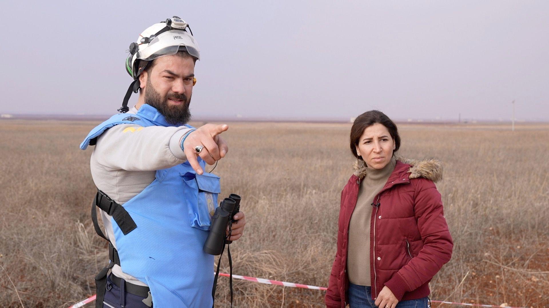 Man wearing PPE - blue flak jacket and a white helmets and holding binoculars. Man points to a location, reporter next to him looking into the distance. 