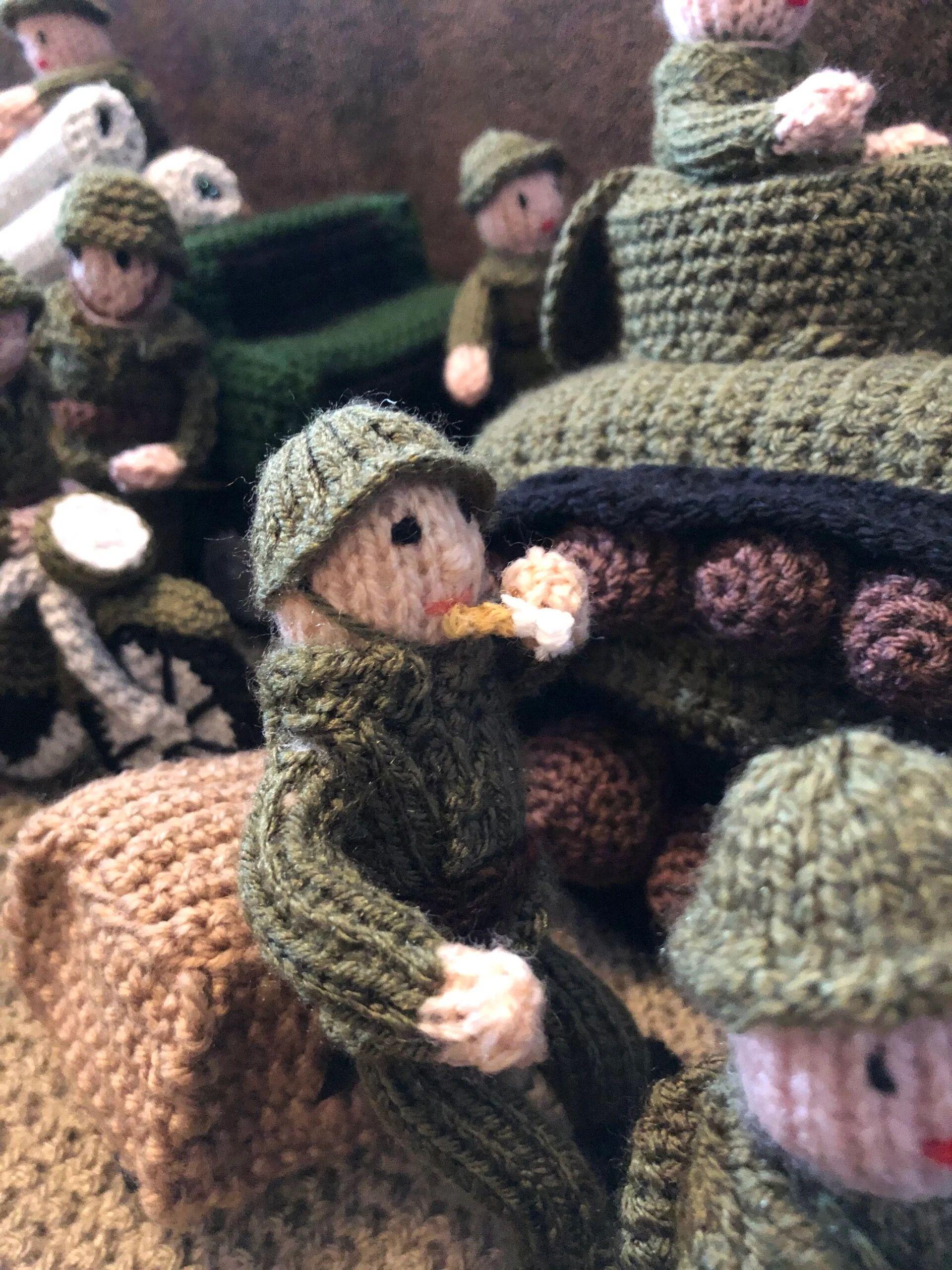 A knitted solider is sitting next to a tank smoking a cigarette. Behind him are more soldiers wearing green outfits and helmets.