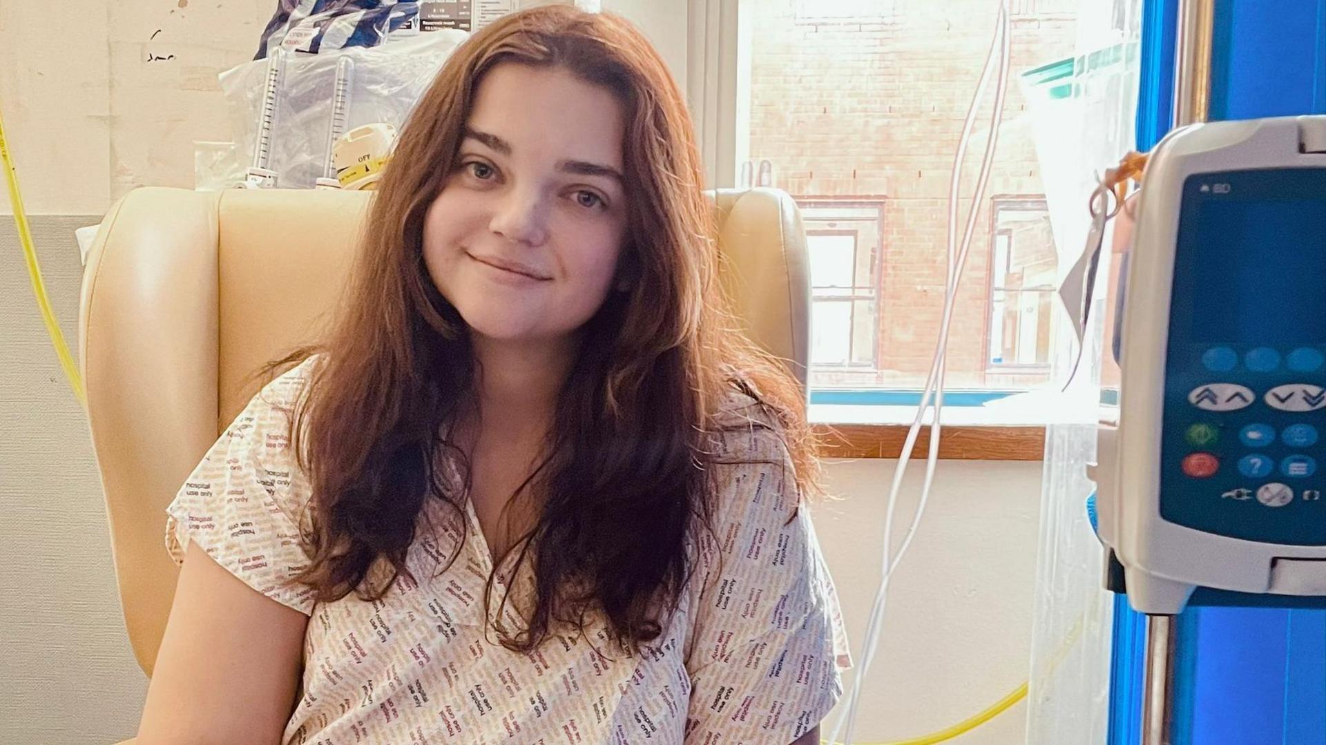 Amy White is sitting on a peach-coloured chair in hospital. She is wearing a hospital gown and is connected to medical equipment.