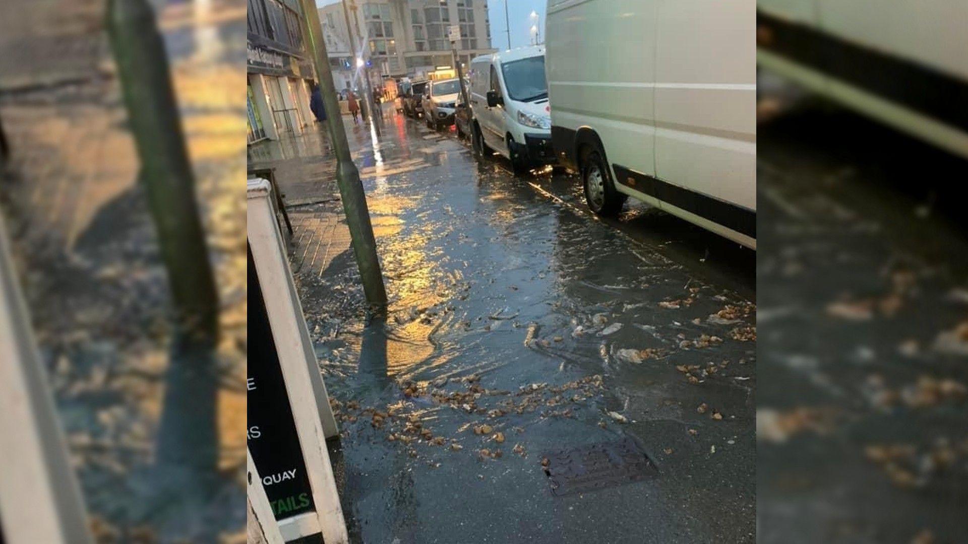 A pavement covered in floodwater on a wet and dark evening. To the right of the image are several parked cars and vans. The water is flowing off the pavement. There are items of debris in the water that appear to be raw sewage.