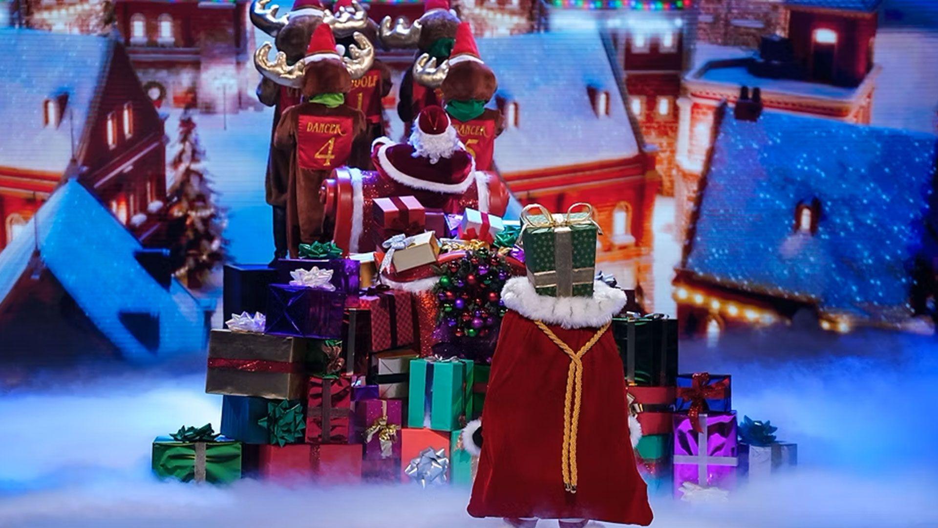A singer dressed as a sack of presents stands next to a pile of presents and dancers dressed as reindeer
