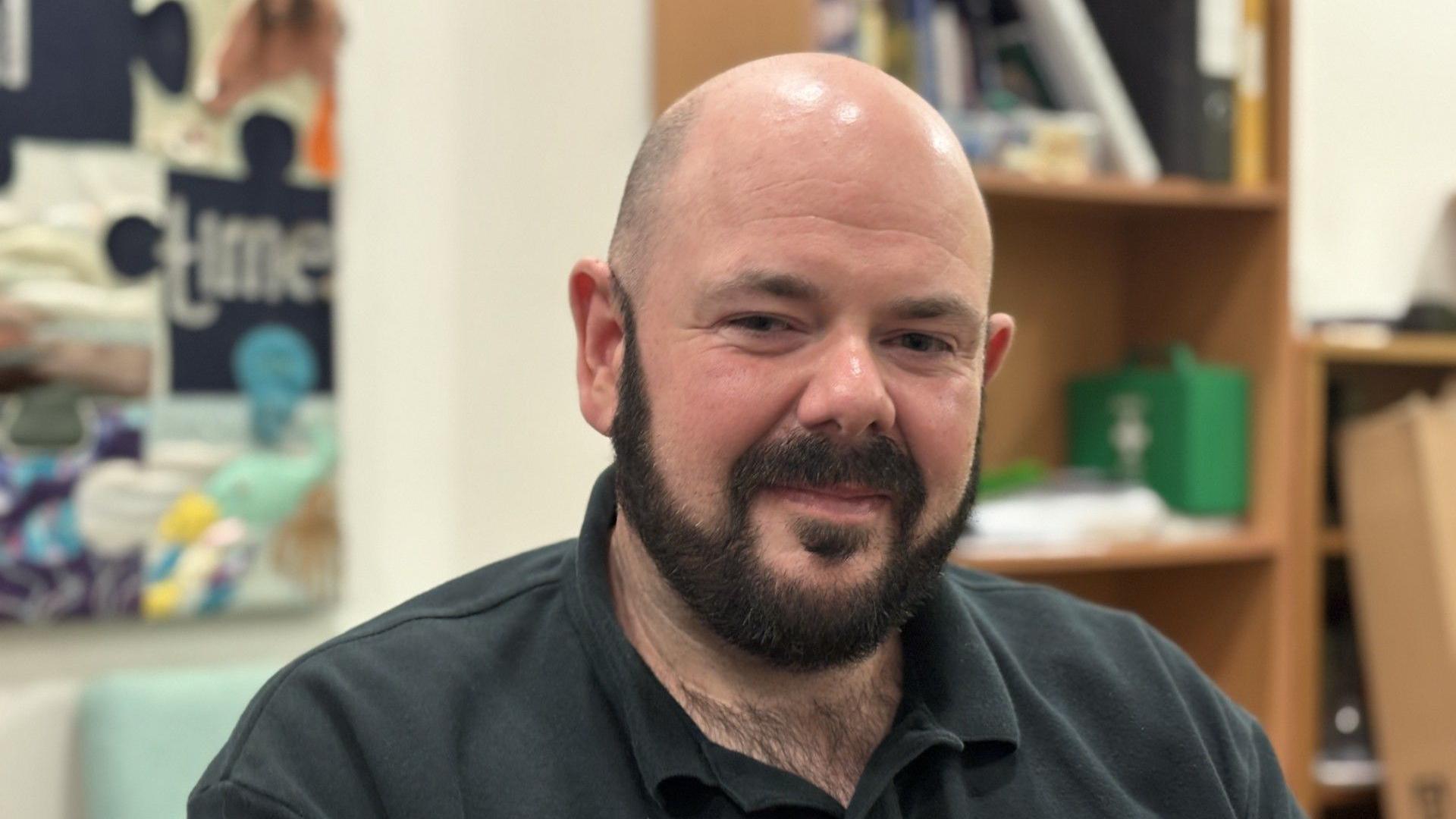 A bald man with a black beard is pictured wearing a black polo top in a classroom environment with shelves and posters behind him. 